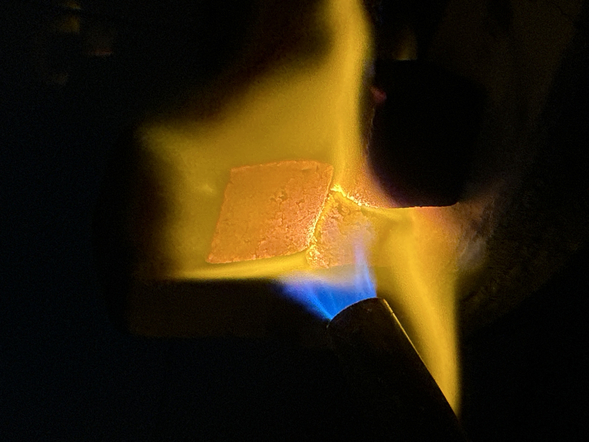 A close-up shot in dim lighting shows a butane torch with a blue flame pointed at a small, light orange, square-ish object. The object is positioned to the left of the flame, which is surrounded by a bright yellow haze. The torch appears to be held by an unseen person. A dark background surrounds the scene. The location of Bac Nha Trang Ward, Vietnam, is not evident from the image.