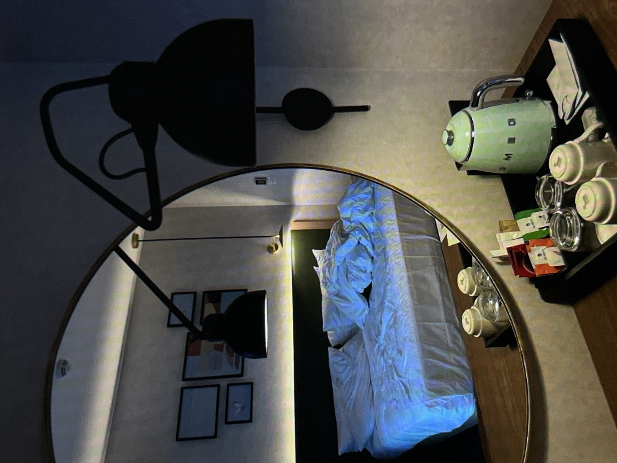 A hotel room is depicted, featuring a bed with rumpled white linens, a headboard, and a circular mirror reflecting the room. The mirror also shows framed artwork on the wall. To the right of the bed, a tray holds a light green electric kettle, cups, and tea or coffee packets. The lighting is dim, with a prominent shadow of a black lamp visible in the mirror. The overall atmosphere suggests a nighttime setting. The specific location within the city of Unknown, Unknown cannot be determined from the visual information provided.
