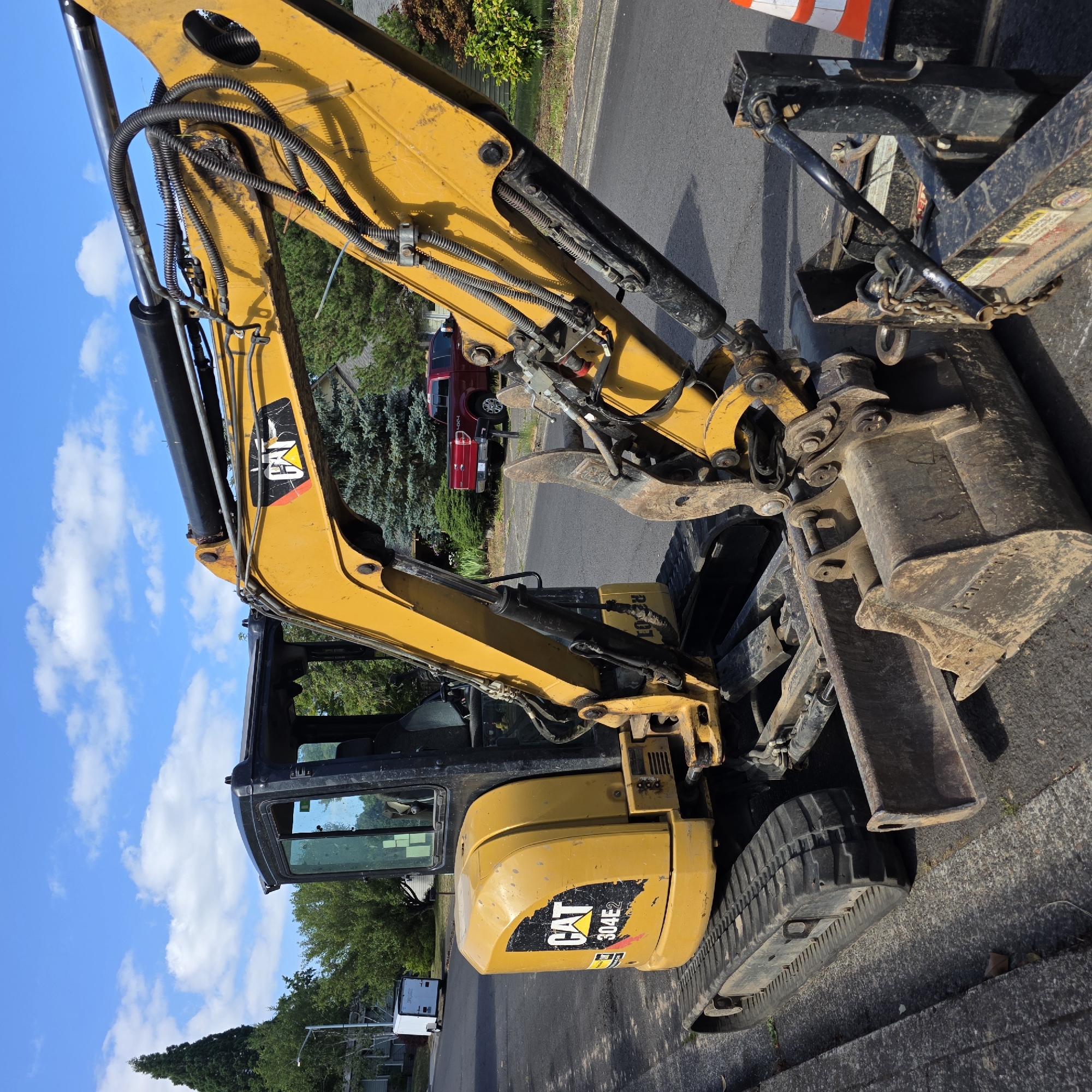An excavator, identified by the "CAT 304E2" logo, is parked on the side of a street in Beaverton, Oregon. The excavator is yellow and black, with tracks and a bucket. It appears to be a construction site, as there are construction cones and other equipment visible in the background. The sky is blue with scattered clouds. A red pickup truck is parked in the background.