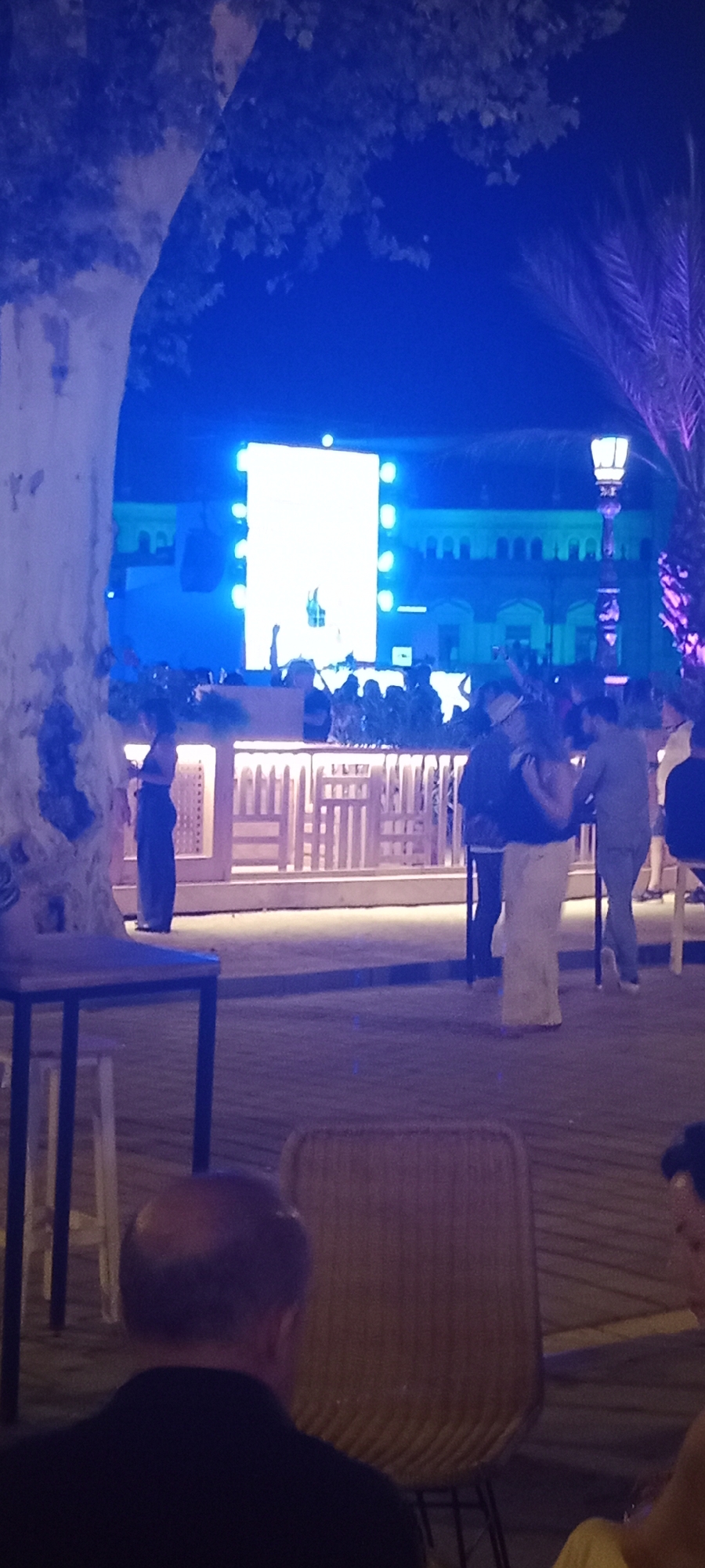 An outdoor night scene in Seville, Spain depicts a vibrant event with a large LED screen broadcasting a performance. The screen is bright white and illuminating the area. A crowd of people are gathered in front of a bar or stage, some raising their hands. One person is visible behind a console, possibly a DJ. A palm tree with purple lighting is visible on the right side of the image. In the foreground, a person is seated at a table, their back to the camera. Wooden chairs and tables are scattered around the venue. The architecture in the background appears to be historical with arches and windows. The overall atmosphere suggests a lively concert or festival.