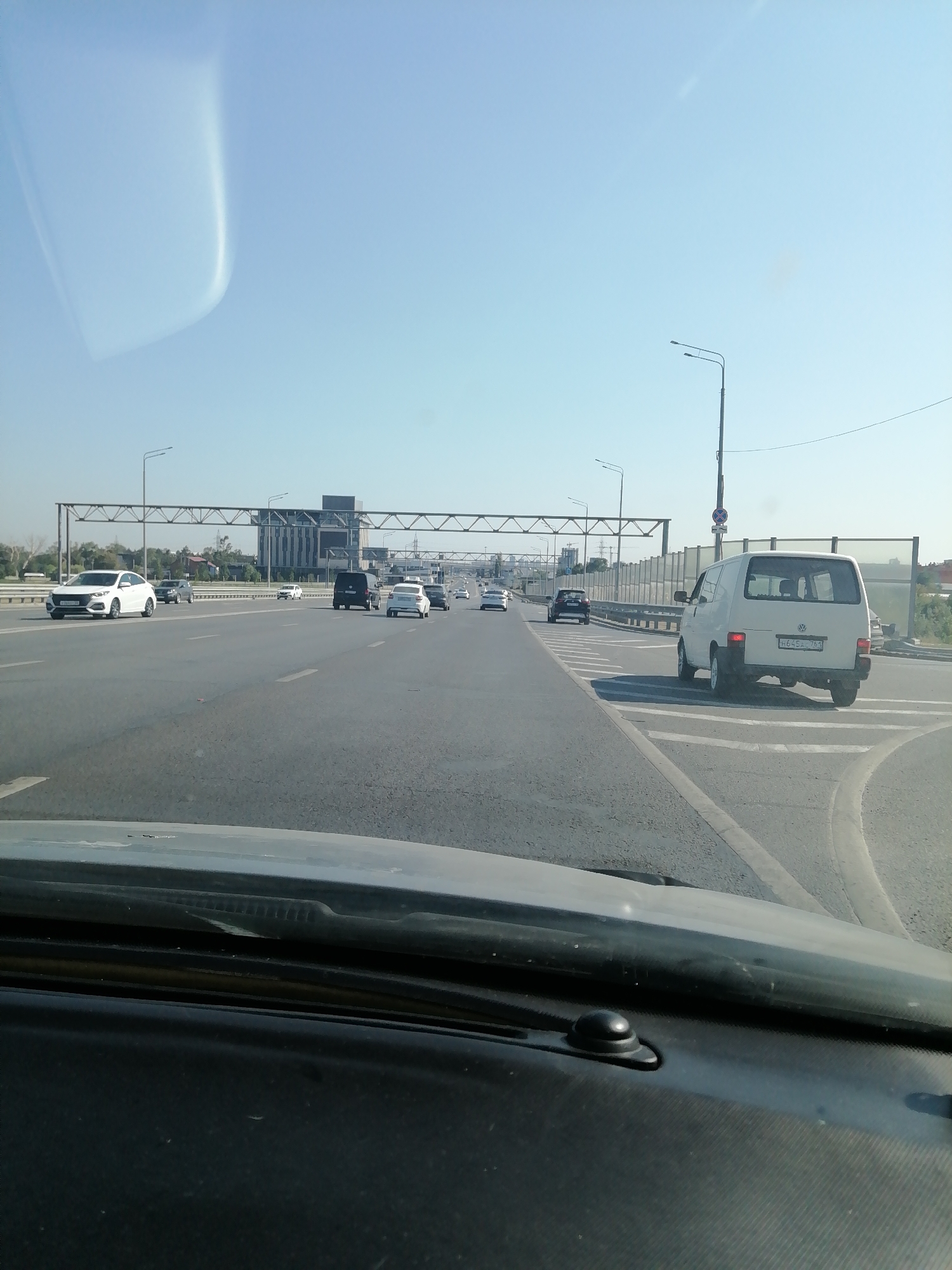 The image depicts a view from inside a vehicle, looking out at a multi-lane highway. The road surface is asphalt with clear lane markings. Several vehicles are visible on the highway, including cars and a white van. In the background, there are large buildings and infrastructure, possibly part of a city or industrial area. The sky is clear and blue, suggesting a sunny day. The foreground shows the interior of the vehicle, including the dashboard and windshield. The license plate of the white van in the right lane reads "H 645 EA 762". The overall impression is a typical urban highway scene.