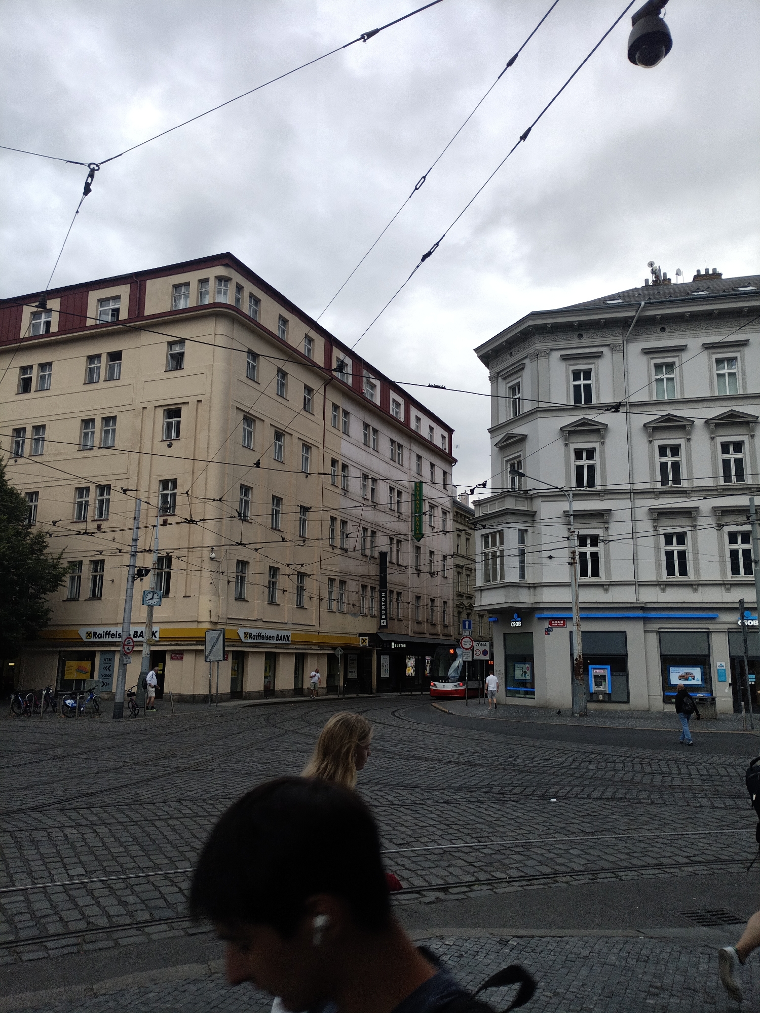 The image depicts a street scene in Prague, Czechia, under an overcast sky. In the foreground, the blurred silhouette of a person's head and shoulders is visible. The midground features a street with tram tracks, paved with cobblestones. A woman with blonde hair is walking across the street. To the left, a cream-colored building with multiple windows and a red-tiled roof dominates the view. The building has a sign for "Raiffeisen BANK." To the right, a lighter-colored building with ornate architectural details and numerous windows stands out. This building also has signage for "CSOB" and "Raiffeisen BANK." A red and white tram is visible turning a corner in the distance. The environment is urban, with power lines crisscrossing the sky above the street. Several bicycles are parked near the cream-colored building.