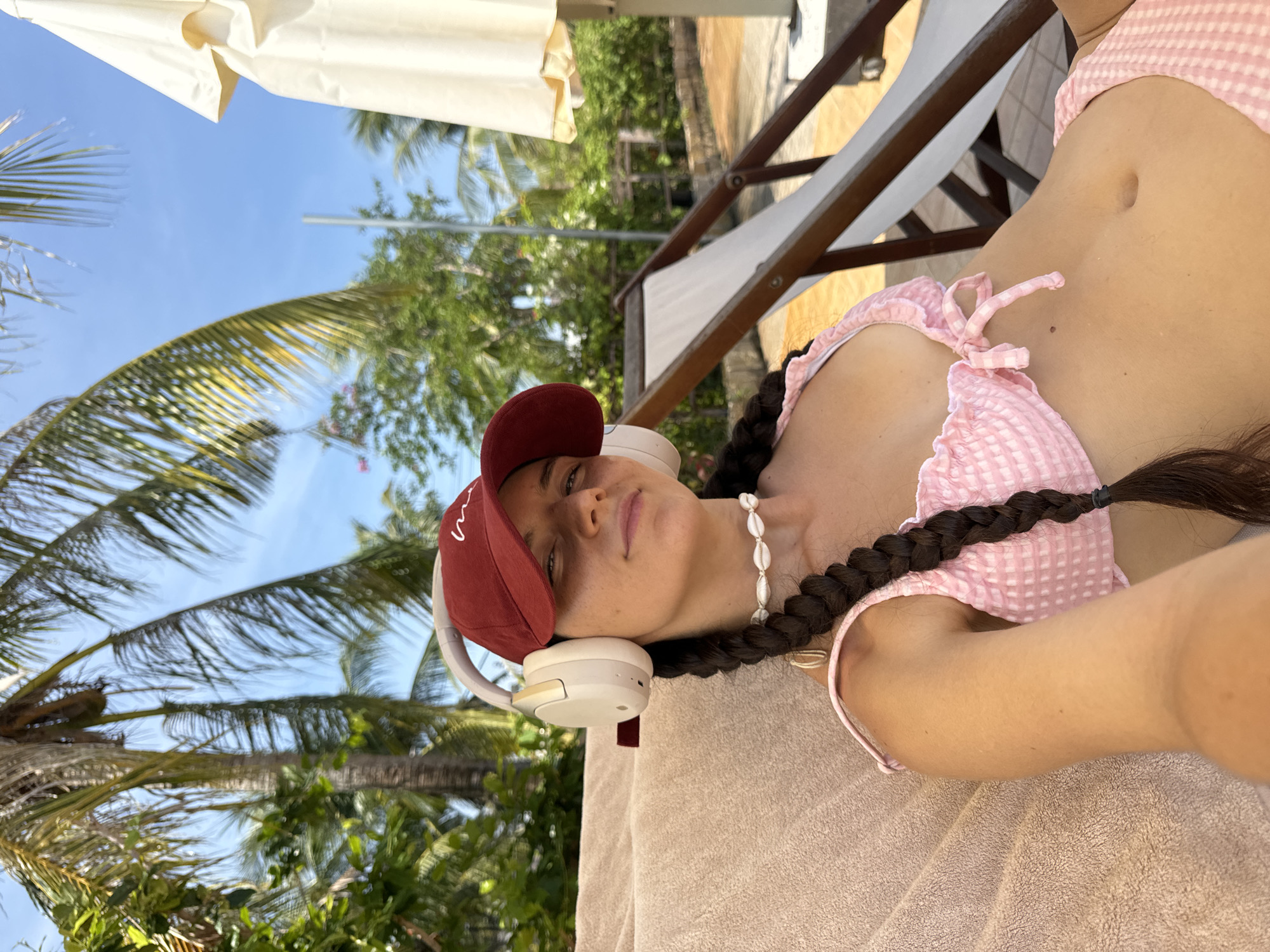 A woman with dark hair in a braid and wearing a red baseball cap and pink gingham bikini is relaxing on a lounge chair under palm trees. She is wearing white headphones and a shell necklace. The background shows more palm trees, lush green foliage, a bright blue sky, and a white umbrella. The scene appears to be a resort or beach area.