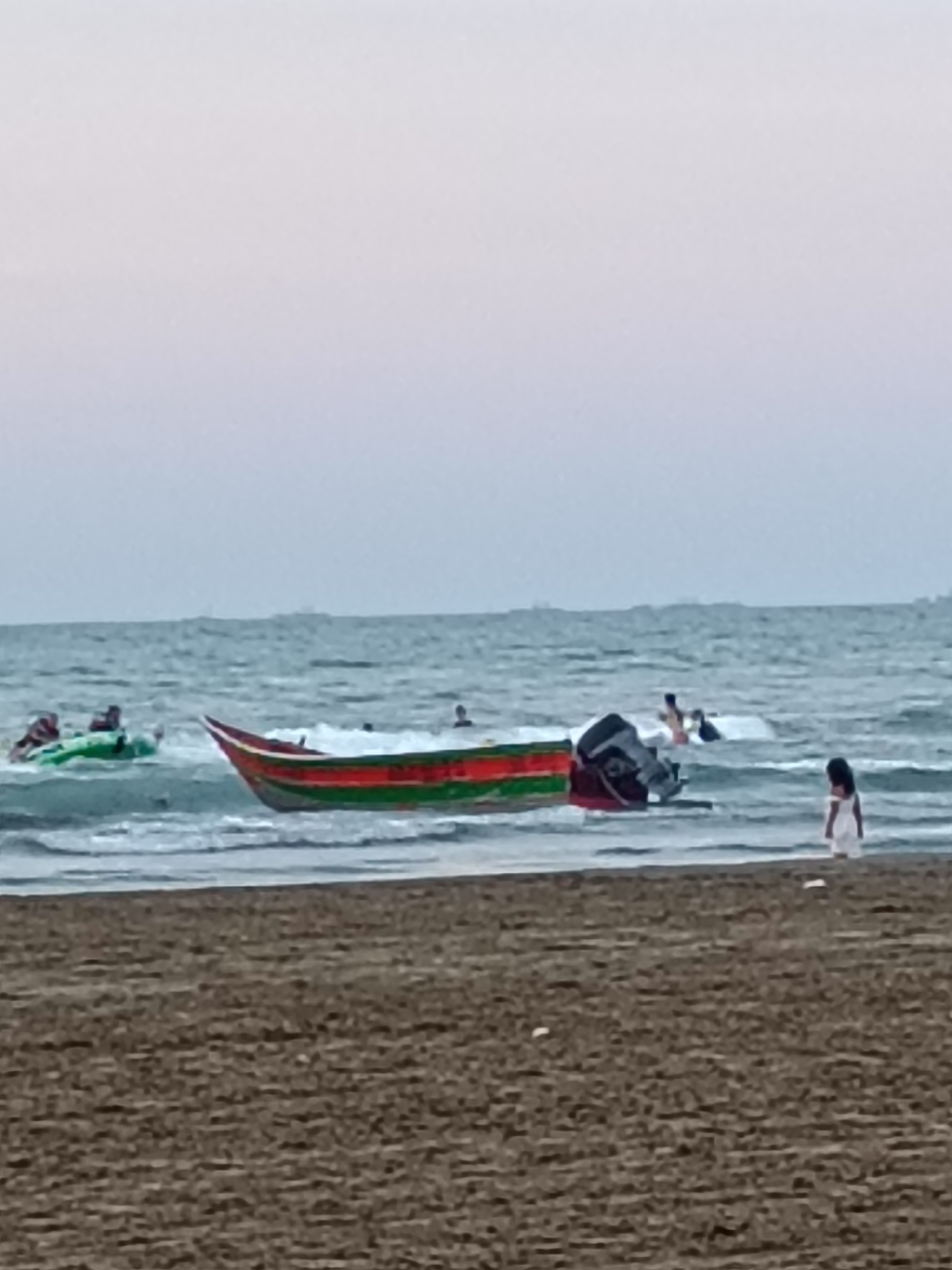 A wide shot of a beach with choppy water and a low horizon under a pale sky shows several people and a boat. In the foreground, a sandy beach stretches across the bottom of the frame. In the middle ground, the ocean is populated by a small, red, green, and white striped boat with an outboard motor at the stern, its bow pointing towards the shore. Further out, a green inflatable raft with at least two people is being pulled through the water by a person in the water. A lone person in a white dress stands on the wet sand at the water's edge on the right side of the frame. In the distance, other indistinct figures are visible in the water. The overall atmosphere is calm, with gentle waves lapping the shore. The scene is set in Khazar Raion, Azerbaijan, a coastal district known for its beaches and recreational activities.
