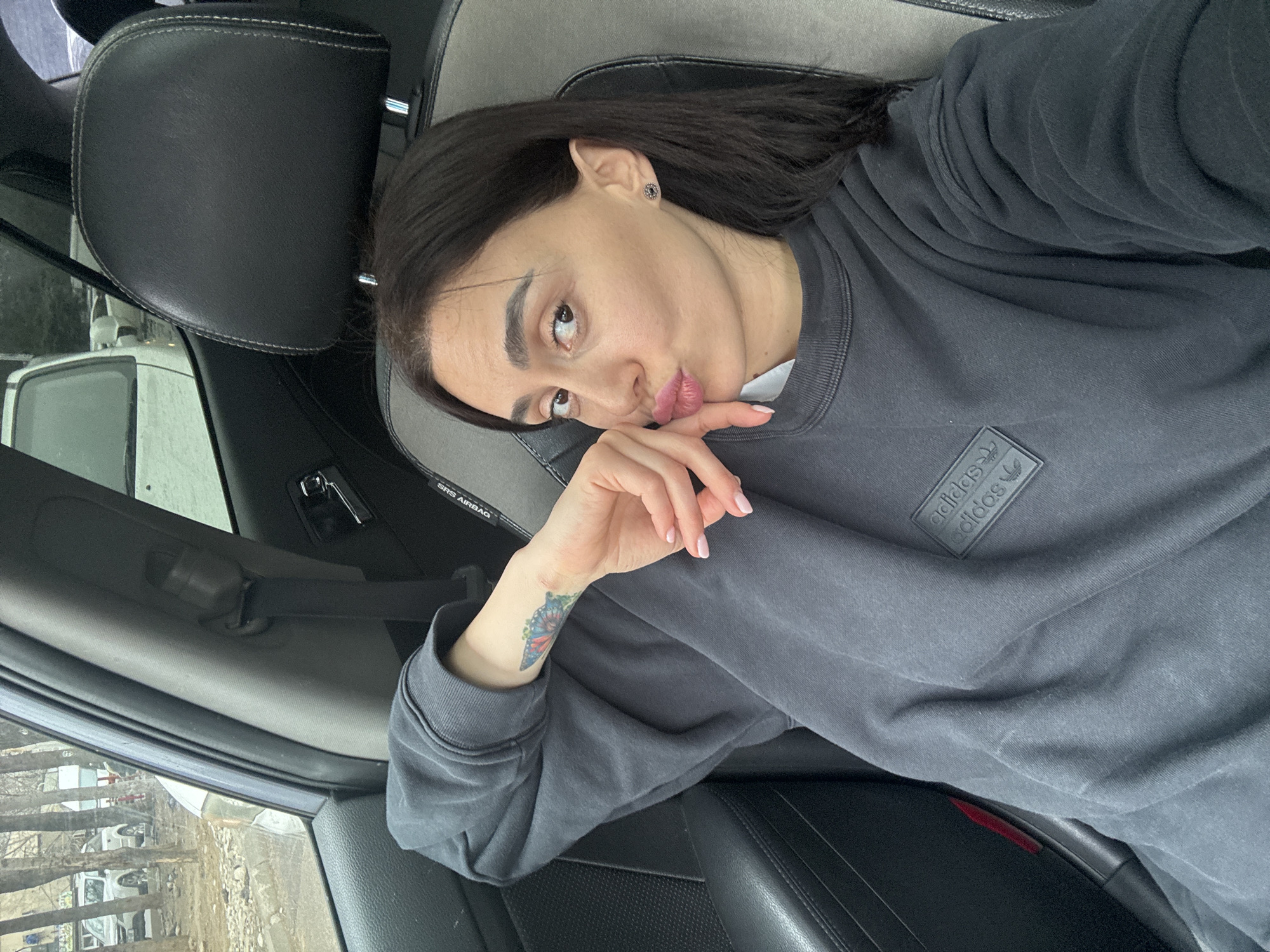 A female individual is seated in the front passenger seat of a vehicle, located in Tbilisi, Georgia. She possesses dark hair, light-colored skin, and dark eyes, looking directly at the camera with her lips pursed in a pouting expression. She wears a dark grey long-sleeved sweatshirt with a black rectangular "adidas" logo patch on the upper left chest. A multi-colored butterfly tattoo is visible on her left forearm, and her fingernails are painted with a light pink polish. Her left hand is positioned with fingers resting near her chin. A dark stud earring is present in her right ear. The car's interior features dark and light grey upholstery on the seats, and a black seatbelt with an "SRS AIRBAG" label is visible on the pillar to her right. Through the passenger side window, an exterior urban or suburban scene is visible, showing bare trees, a paved surface, and several other vehicles, including a white SUV, under what appears to be an overcast sky.