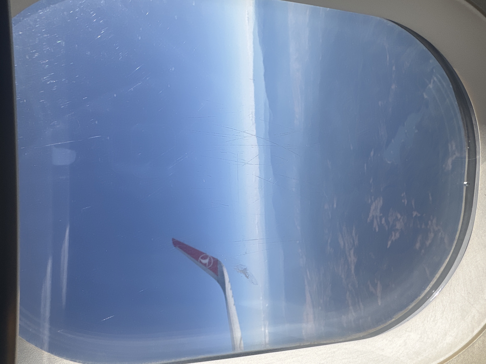 An aerial view from an airplane window shows a clear blue sky and distant mountains. A portion of the airplane's wing, colored white with a red tip and the Turkish Airlines logo, is visible on the left side of the frame. The window has visible scratches and smudges. Below the mountains, a landscape of varied terrain with hints of vegetation and open spaces can be seen. The image is taken during daylight hours.