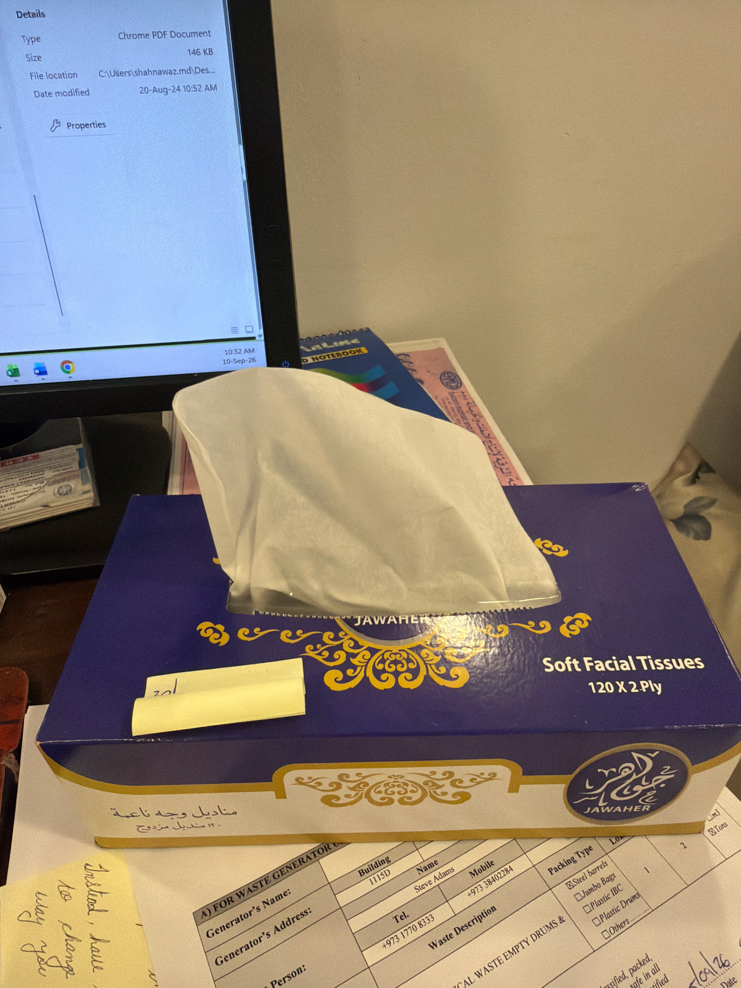 The image depicts a desk scene, with a focus on a box of "JAWAHER Soft Facial Tissues" with Arabic script. A white tissue is partially pulled out of the box. To the left of the tissue box, a computer monitor displays file details, showing "Chrome PDF Document," size "146 KB," file location "C:\Users\shahnawaz.md\Des...", and date modified "20-Aug-24 10:52 AM." The bottom right of the screen shows the time as "10:32 AM" and date as "10-Sep-26". A form labeled "A) FOR WASTE GENERATOR" is visible beneath the tissue box, with fields for Generator's Name, Generator's Address, Building, Name, Mobile, Tel., Waste Description, and Packing Type. Some entries on the form are visible, including "Steve Adams," "+973 38402284," and "+973 1770 8333." There are also handwritten notes on yellow sticky papers. The overall environment suggests an office or workspace.