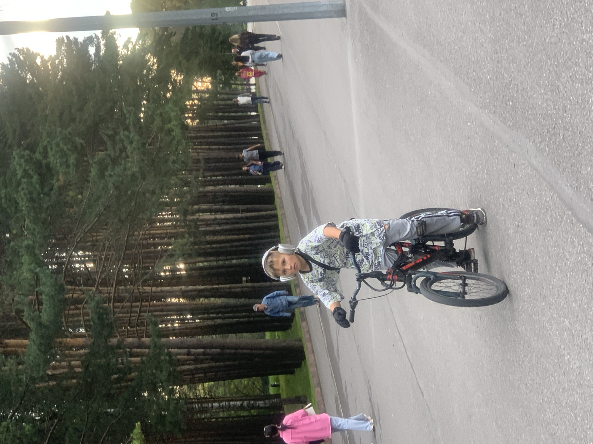 The image shows a boy on a bicycle in what appears to be a park in Prokopyevsk, Russia. The boy is wearing headphones, a patterned shirt, grey pants with a stripe, and gloves. He is riding on an asphalt path lined with tall trees. Several other people are walking in the background on the same path. The sun is setting, casting a warm glow through the trees. A tall, gray pole with the number "19" is visible on the right side of the image.