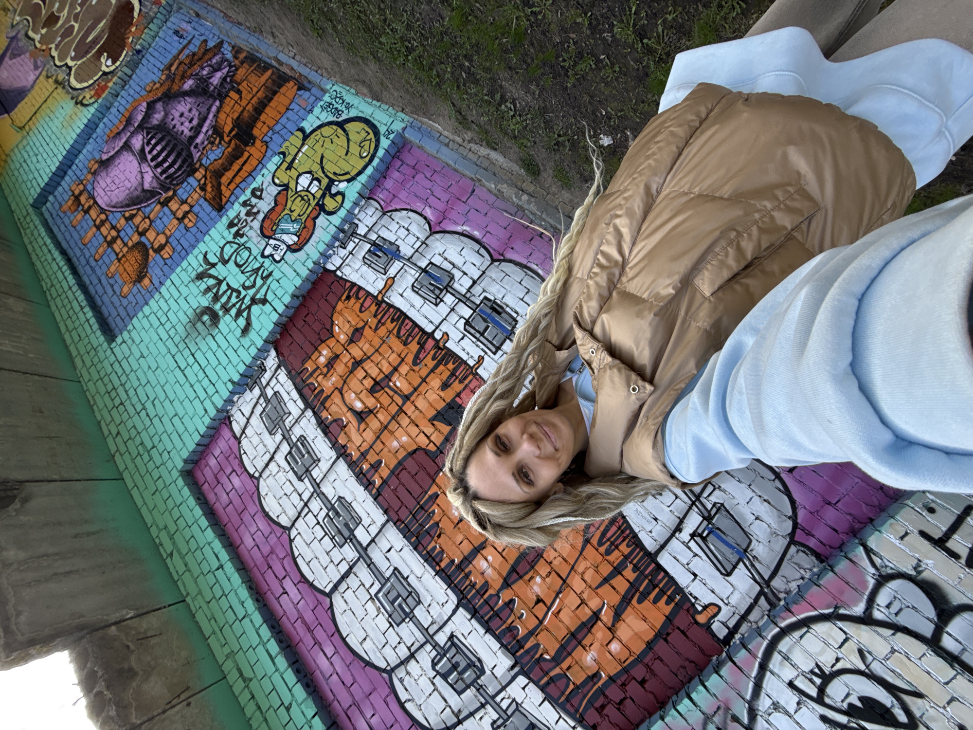 A young woman with blonde braided hair smiles at the camera while lying on a graffiti-covered brick wall. She wears a light blue sweatshirt and a light brown puffer vest. The brick wall features colorful graffiti art, including a stylized purple monster-like character, a yellow cartoon figure, and abstract patterns and text on a background of green, purple, orange, and white bricks. The location appears to be a mural or street art installation, possibly in an urban area. The presence of the graffiti art suggests the setting may be located in Rusanovo, Russia.