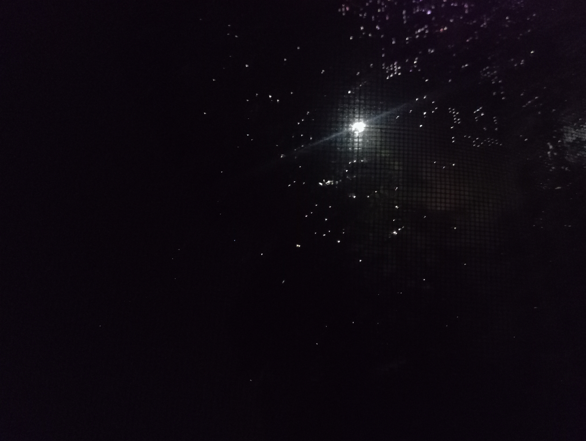 A dark scene, likely viewed through a window with a screen. Water droplets are visible on the windowpane, reflecting light. A distant light source, possibly the moon or a street lamp, is visible through the screen. The location, Nkpor, Nigeria, cannot be verified from the image alone. No people, specific subjects, objects, actions or interactions are discernable beyond the aforementioned details. The environment appears to be an outdoor, nighttime setting.