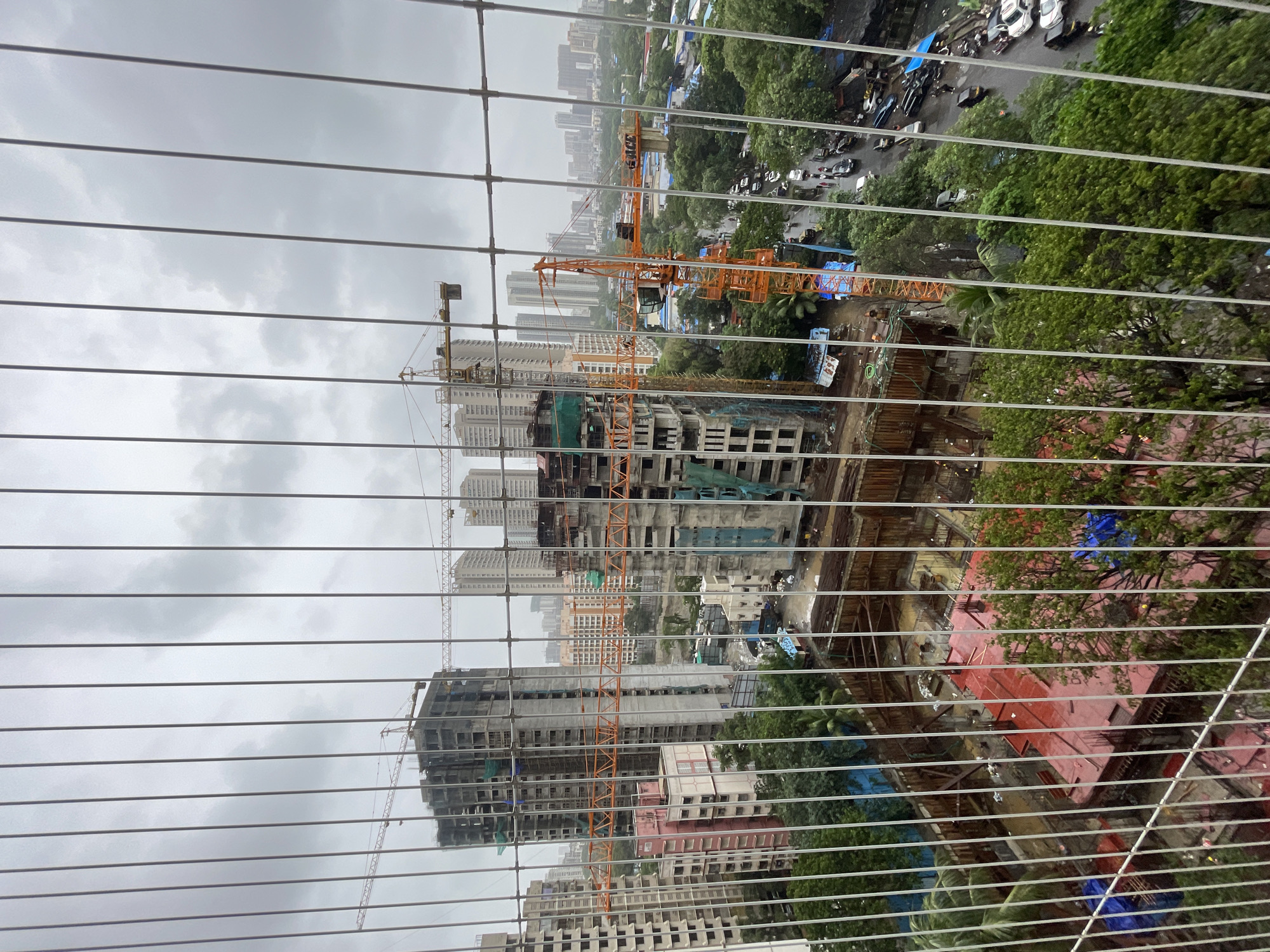 This is a high-angle, medium-shot, exterior view of a busy construction site in Mumbai, India, as seen through a wire mesh. The scene is dominated by several unfinished high-rise buildings and construction cranes, indicating a developing urban area. A prominent orange tower crane is positioned in the foreground, with its jib extended over the construction site. To its left, another, smaller crane is visible. The buildings under construction are multi-story structures with exposed concrete frames and scaffolding. Some buildings appear to be clad in light-colored panels, while others are still in their raw concrete stage, with green netting draped over parts of their exteriors.
In the foreground, a deep excavation pit is visible, revealing a red-colored foundation with visible rebar. Several trees, some with lush green foliage, surround the construction site and extend into the middle ground, providing a contrast between the natural and the built environment. Beyond the immediate construction zone, a dense urban landscape unfolds, with numerous residential and commercial buildings stretching towards the horizon. The sky is overcast with gray clouds, suggesting a cloudy or potentially rainy day. A street runs through the middle ground, with several vehicles, including cars and what appear to be auto-rickshaws, navigating the road. A few individuals are discernible on the street and near the construction site, engaged in activities typical of such an environment. The overall impression is one of ongoing development and urban expansion in a densely populated city.