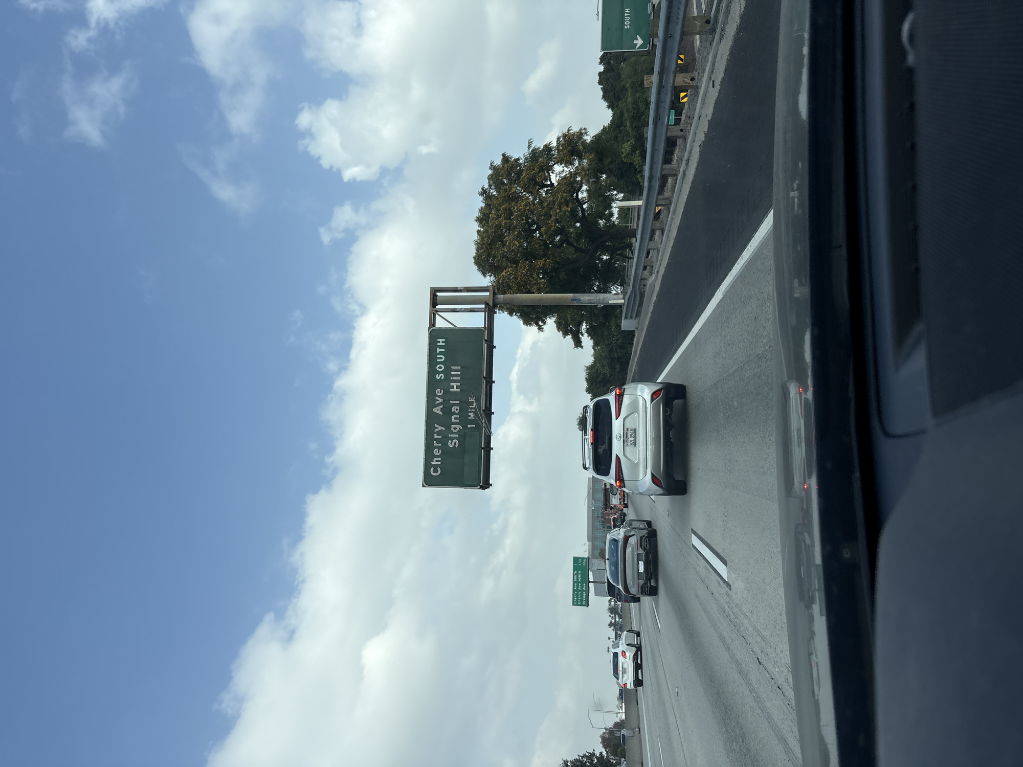The image captures a view from a vehicle on a highway in Long Beach, California. A large green overhead sign indicates the Cherry Ave South/Signal Hill exit is 1 mile ahead. Other vehicles, including a white SUV, are visible in the adjacent lanes. The sky is partly cloudy. The road appears to be a multi-lane highway with a concrete barrier on the right side. Vegetation, including trees, can be seen beyond the barrier. The image is taken from within a vehicle, as evidenced by the car frame visible on the right side.