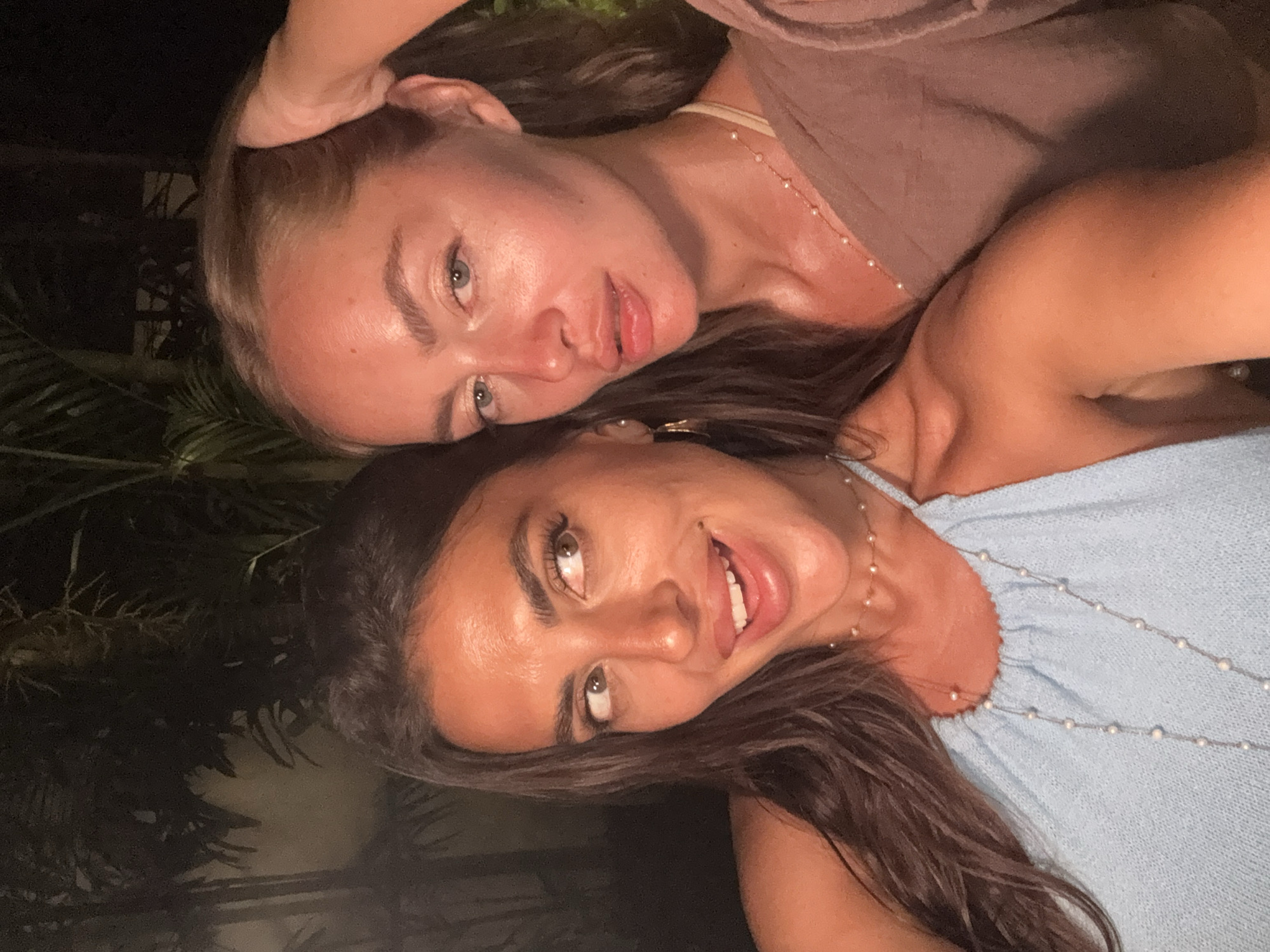 Two women are closely posed, possibly for a selfie. One woman has light skin, blonde hair, and is wearing a tan-colored top. The other woman has tan skin, brown hair, and is wearing a light blue top. Both appear to be wearing necklaces. The background is dark, with some foliage visible, possibly indicating an outdoor setting. Given the presence of tropical vegetation, the image may have been taken in Kuta Selatan, Indonesia.