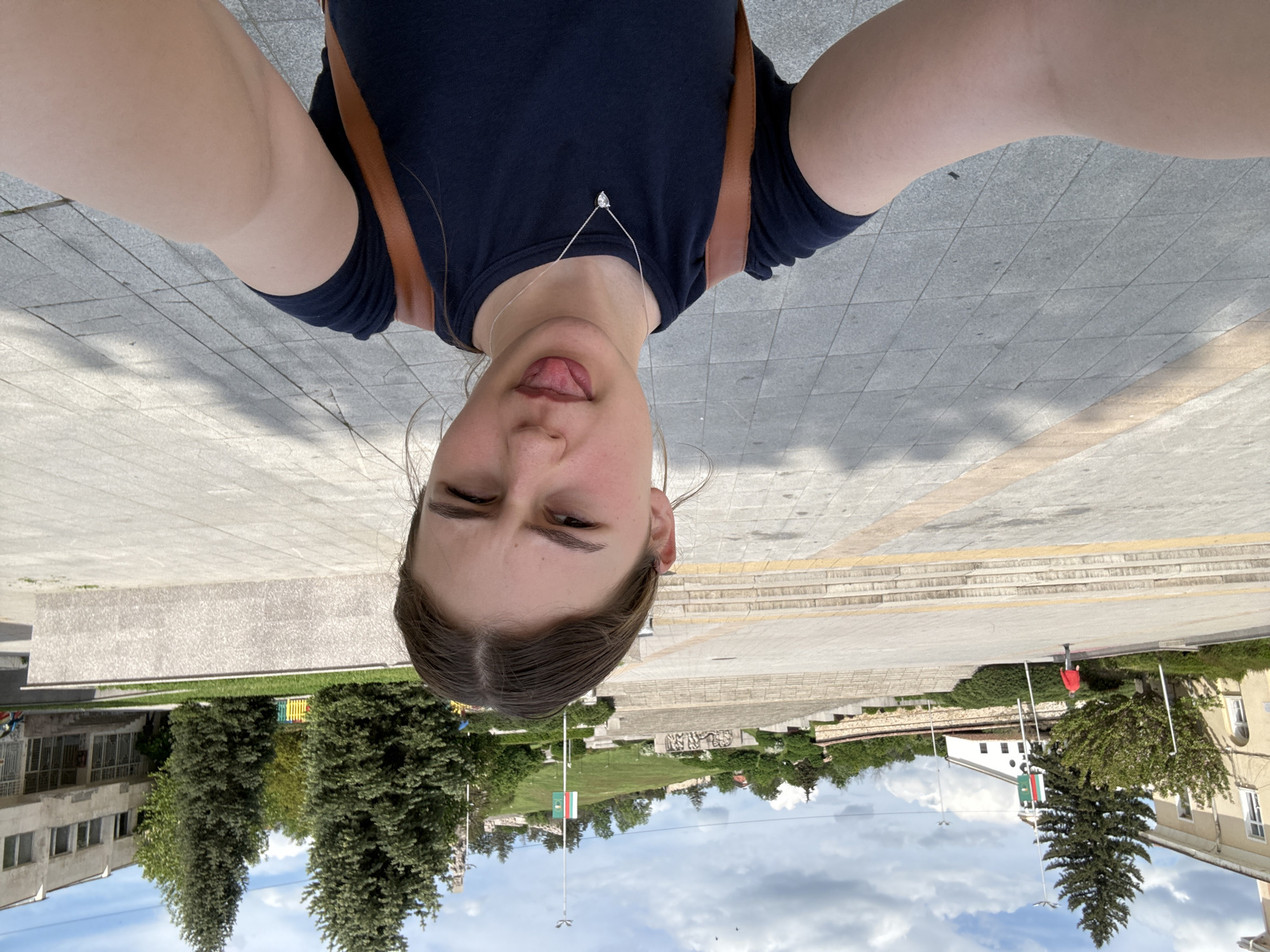 A selfie taken with the phone held upside down, shows a young woman with her tongue sticking out. She is wearing a navy shirt and a silver necklace. Behind her is the city of Panagyurishte, Bulgaria, including the monument complex, the Bulgarian flag flying on a pole and other buildings with green trees. The sky is cloudy. In the distance, a person wearing red can be seen. The ground she is standing on is paved with square stones. She also has a backpack.