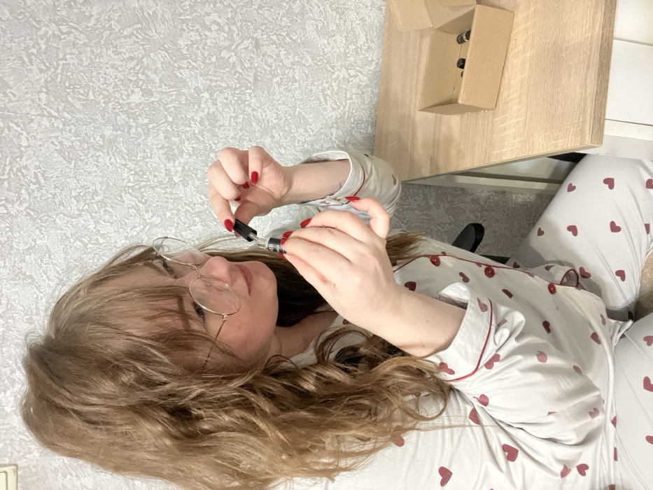 A fair-skinned female, with long, wavy light-brown hair pulled into a high ponytail and bangs across her forehead, is depicted in a residential setting in Chaikovsky, Russia. She wears round, thin-rimmed glasses and light-colored pajamas patterned with red hearts, featuring red piping and red buttons. Her fingernails are painted red. She is seated at a light-colored wooden desk, holding a small, dark-colored glass bottle with her left hand, while her right hand operates a black dropper cap inserted into the bottle, suggesting either extraction or dispensing of liquid. A partial label on the bottle is visible, including the letters "CBD". To her right, on the desk surface, is an open, light-brown cardboard box containing multiple identical small, dark-colored bottles. The background wall is covered with a light grey, textured wallpaper.