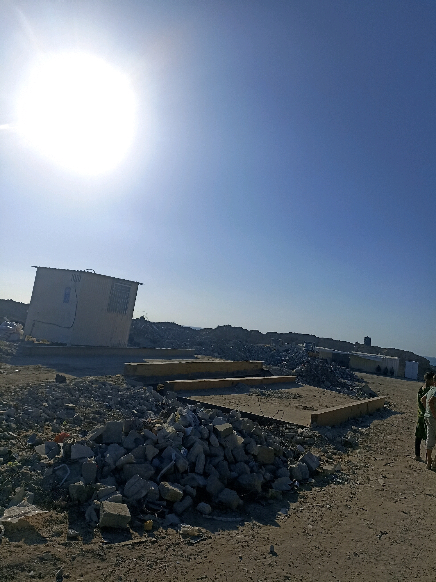 A wide-angle shot captures a sunlit, clear blue sky over a devastated landscape in Gaza. The foreground is dominated by piles of rubble, including large concrete chunks and scattered debris. A single, pale-colored corrugated metal structure, possibly a portable office or shelter, stands slightly tilted on a concrete foundation in the mid-ground to the left. Behind this structure, more rubble and debris extend into the background, interspersed with several construction containers or similar modular units. A faint outline of the sea is visible on the horizon. On the right side of the frame, three individuals stand in the distance, partially obscured by the debris and sunlight. One appears to be wearing a green shirt and dark shorts, while another is in a white shirt and light-colored shorts. The overall scene depicts destruction and reconstruction, characteristic of areas impacted by conflict or extensive development.