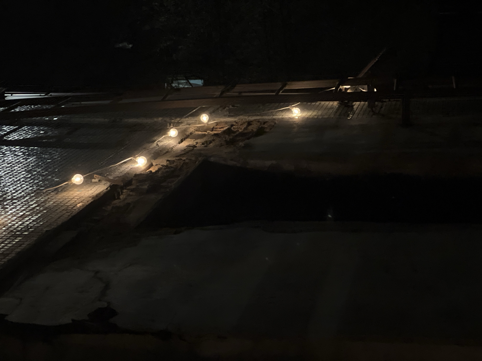 The image depicts a nighttime scene in the city of Mytishchi, Russia, focusing on the exterior of a building. A distressed, light-colored concrete or stucco wall occupies the left and central foreground, exhibiting significant peeling and deterioration that exposes underlying dark brickwork in multiple areas. A dark, rectangular recess or doorway is present on the lower left portion of this wall. Seven warm-toned, spherical light bulbs are strung horizontally across the upper section of the wall, providing localized illumination. To the right of the illuminated wall, a dark vertical metal structure, possibly a fire escape or access ladder, is visible, extending upwards. Above the string lights and to the right, a dark, grid-patterned surface is present on the building's facade. The background is predominantly dark, with indistinct dark foliage visible on the far right and a faint, rectangular light source discernible within a distant opening or window. No individuals, actions, or specific interactions are observed.