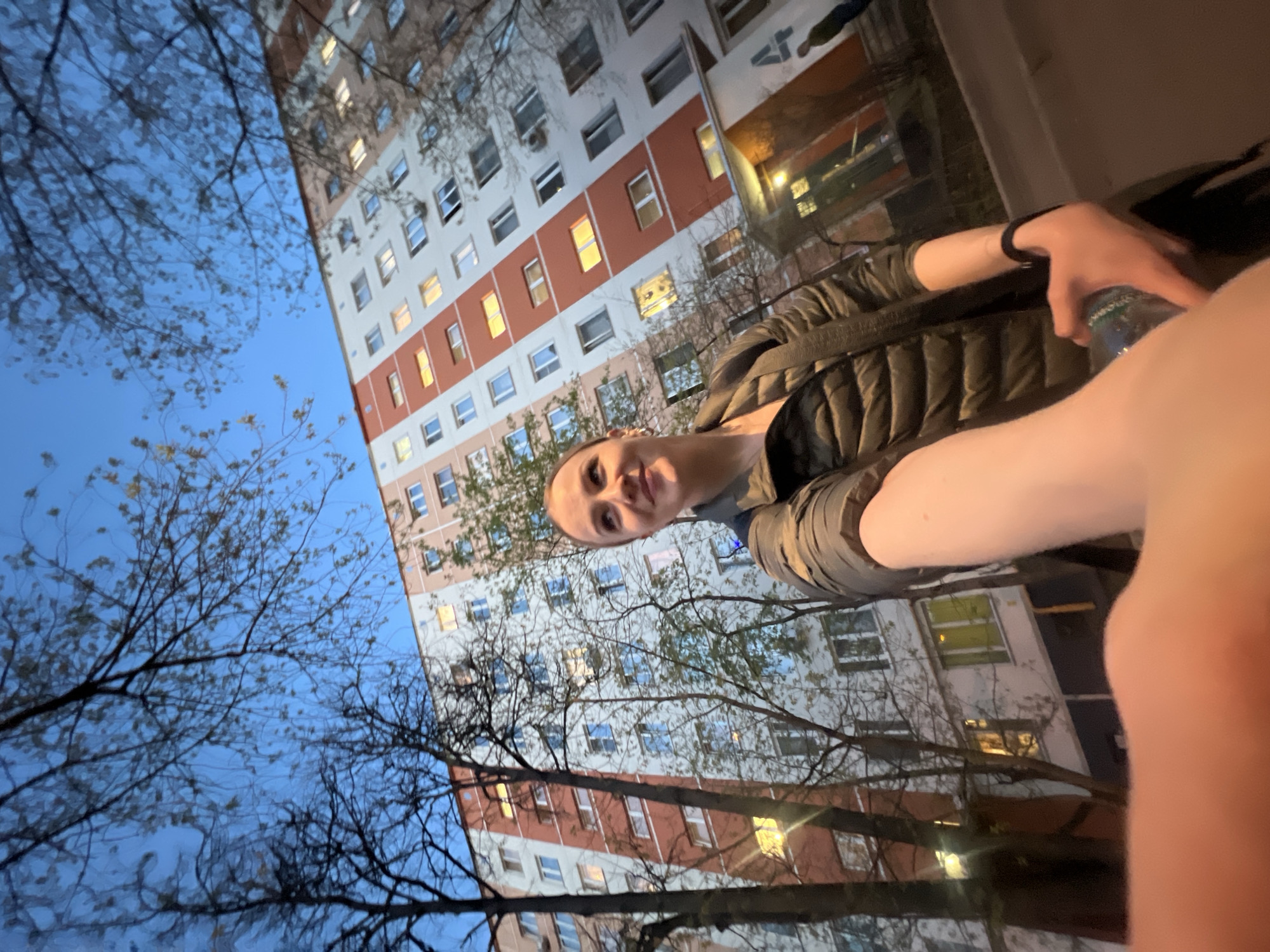 A low-angle, rotated, outdoor shot taken at dusk depicts a fair-skinned person, likely a woman, in the foreground holding a water bottle. She is wearing a dark puffer jacket. Behind her, a tall, white apartment building with rows of windows is visible. Some windows are illuminated. Bare tree branches are silhouetted against a blue sky in the left and bottom portions of the frame. The location may be Khimki, Russia, given that information was provided.