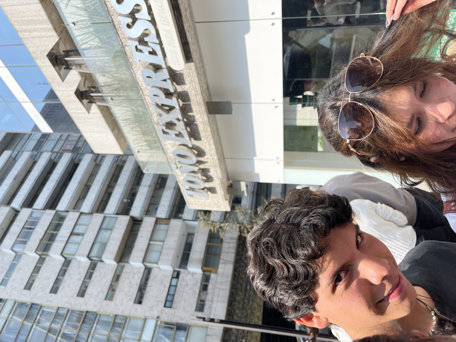 A sideways snapshot shows two people, one wearing sunglasses, in front of a building labeled "TOKYO EXPRESS". The building is tall and constructed from light-colored stone or concrete with a repeating pattern of narrow windows. The two people are close to the camera, only partially visible. The person in the foreground has curly, dark hair. The lighting is bright, suggesting a sunny day. The photograph appears tilted, giving a distorted perspective.