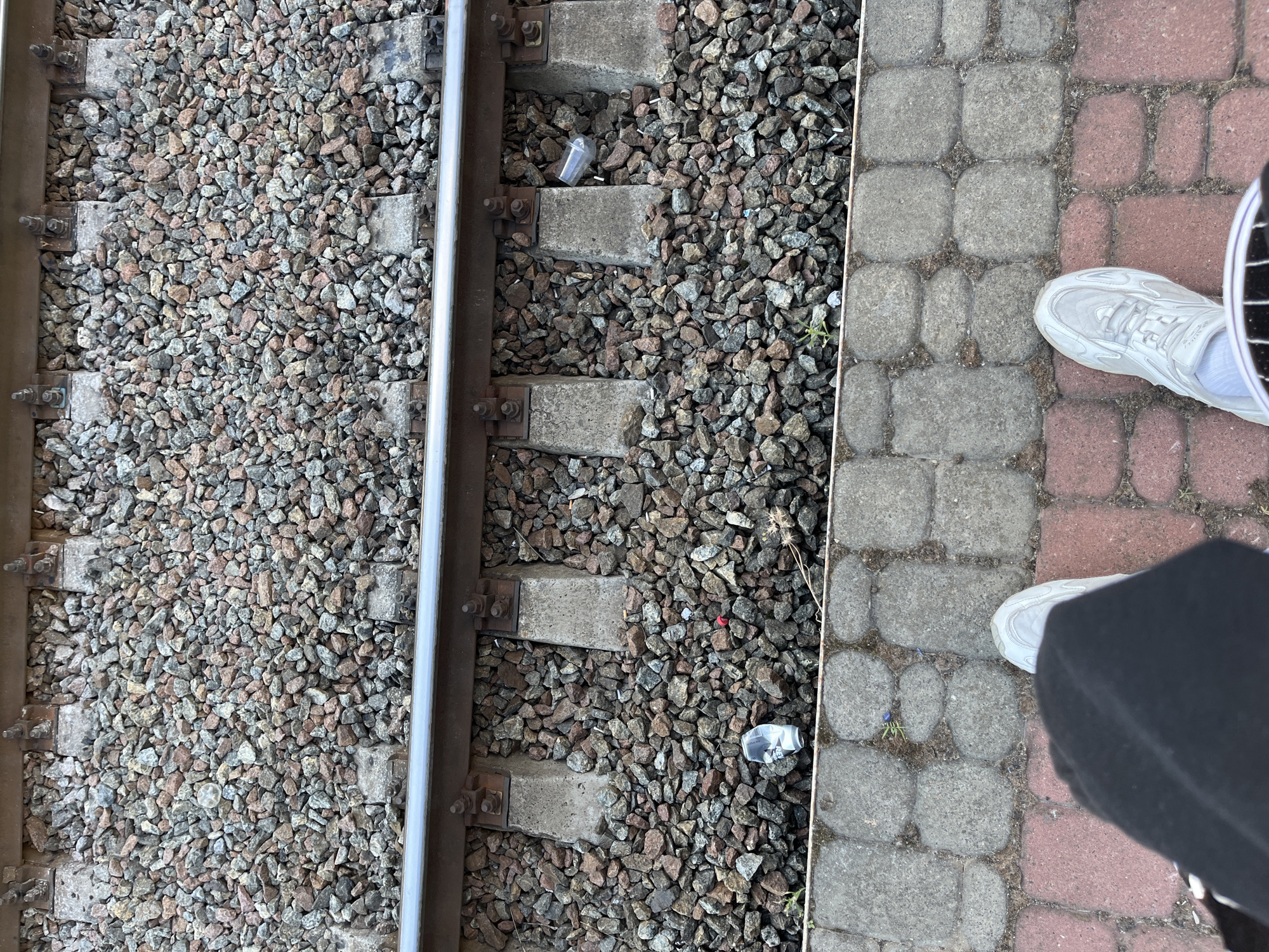 The image shows a section of railway tracks at a station in Kyiv, Ukraine. The tracks are made of steel rails supported by concrete sleepers embedded in a bed of gravel. Several discarded plastic cups and other small pieces of litter are scattered amongst the gravel. In the foreground, a person's feet wearing white sneakers and dark pants are visible, standing on a paved platform. The platform is made of interlocking red and grey bricks. The overall scene suggests a public area that is not well-maintained.