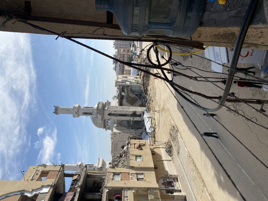 The image depicts a devastated urban street scene in Khan Yunis, Palestinian Territory, under a clear blue sky with scattered white clouds. The perspective is from an elevated position, looking down onto a street and damaged buildings.
In the center-background, the severely damaged Great Mosque of Khan Yunis is prominent, featuring two minarets and a central dome, with significant structural collapse visible in its walls and surrounding areas. Piles of rubble and debris surround the mosque. To the left, a multi-story residential building shows extensive damage, with missing walls, exposed interiors, and laundry hanging from balconies. At its base, a commercial storefront features a dark sign with "SQUID GAME" and other Arabic text. Adjacent to this building are further piles of rubble.
The paved street runs diagonally across the midground. Several individuals are present: one person in a dark green uniform walks left-to-right across the street, followed by a child running. Further down the street, near the mosque, another person stands, and multiple figures are visible in the distance. Shadows are cast across the street from the buildings.
In the immediate foreground on the right, a segment of a light-colored building structure is visible, along with a large, clear plastic water bottle. Numerous black electrical wires and cables, some secured with ties, are strung haphazardly across the right side of the frame, accompanied by a yellow hose or cable. Below these wires, partially obscured figures are visible near a white temporary structure or tent displaying a red crescent logo and Arabic text, suggesting an aid or medical presence.