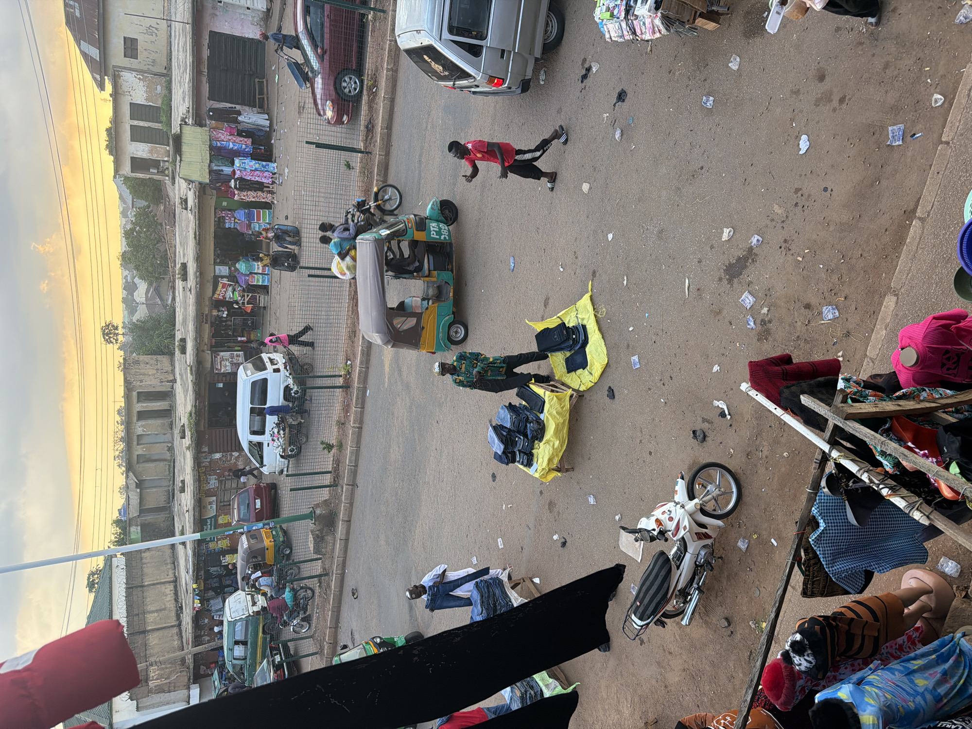 This is a high-angle, vertical shot of a bustling street market in Bukuru, Nigeria, likely taken in the late afternoon or early evening, as indicated by the warm, soft lighting of the sunset in the background. The scene is dynamic, filled with activity and a variety of objects.
Dominating the foreground and midground are several vehicles including a white van, a maroon car, an auto-rickshaw, and multiple motorcycles, some of which are parked and others potentially in use. There are also several people present, some interacting with each other, some tending to wares, and others walking or standing. One person in a red shirt and black pants is striking a pose in the street, giving a thumbs-up. Another person in a patterned shirt and dark pants appears to be arranging items on a yellow tarp on the ground. Scattered throughout the street are items of clothing, including stacks of jeans, and other merchandise.
The setting appears to be a street in a commercial area with buildings lining one side. Some buildings show signs of wear and tear, with peeling paint. Merchandise, including colorful textiles and clothing, is displayed on racks and laid out on the ground, typical of an open-air market. There are also plastic bags and various types of trash scattered on the pavement, suggesting a busy and well-trafficked area.
Notable details include the presence of mannequins dressed in clothing near a makeshift stall. The overall atmosphere seems lively and active, indicative of a typical market day. Visible text includes "PTA 202" on the side of the auto-rickshaw and some unreadable signs on shopfronts. The weather appears to be clear, with a warm glow from the sunset.