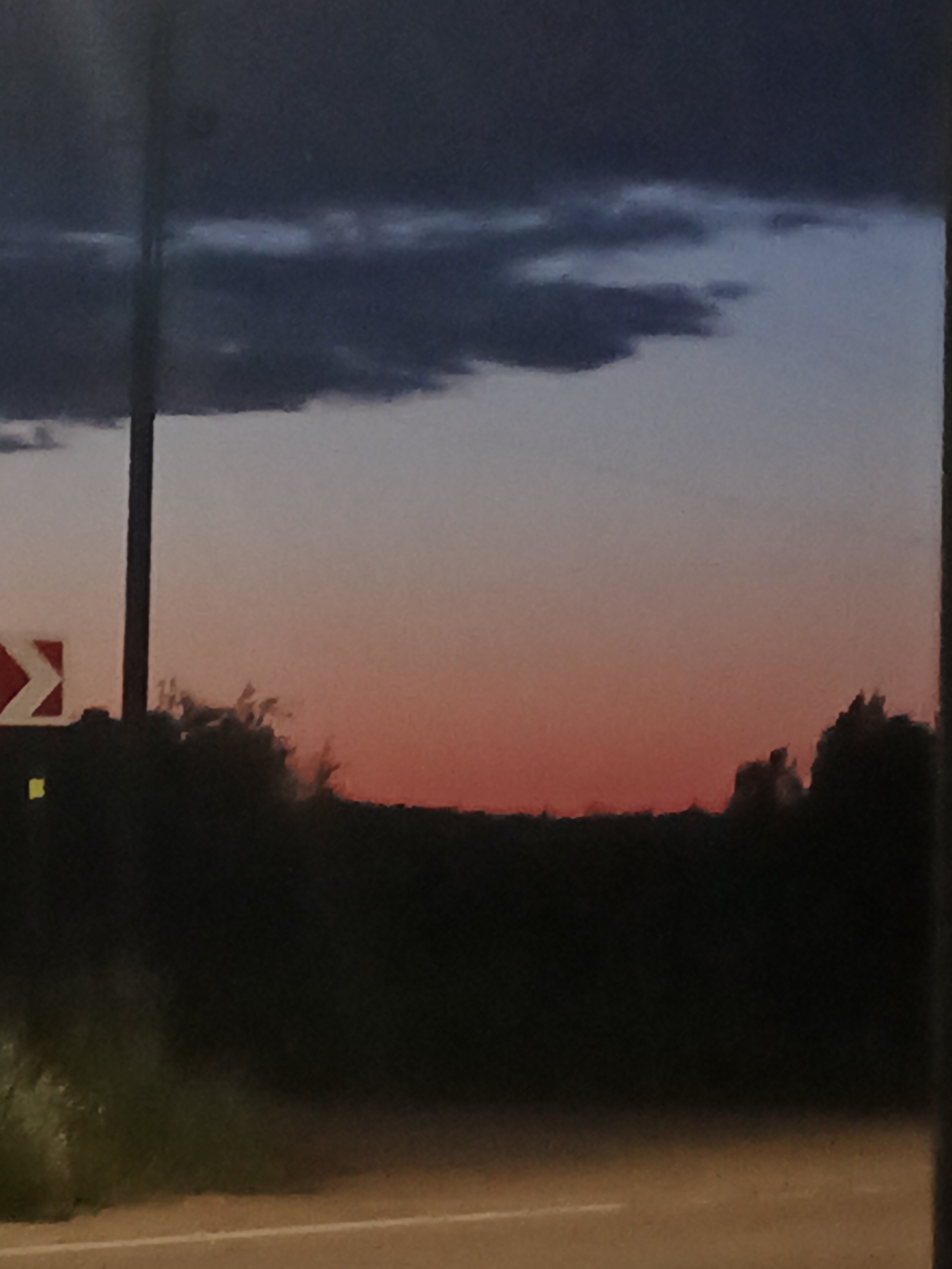 This image captures a twilight scene, likely on the outskirts of Воскресенское, Russia. A dark silhouette of trees and foliage forms the horizon line, against a sky transitioning from a deep blue at the top to a vibrant orange-red near the horizon. Dark, heavy clouds are visible in the upper portion of the sky. In the foreground, a dark road surface with a white line is visible. To the left, a traffic sign with a white arrow on a red background is partially visible, mounted on a dark pole. The overall impression is of a natural landscape under a dramatic sky during the evening transition.