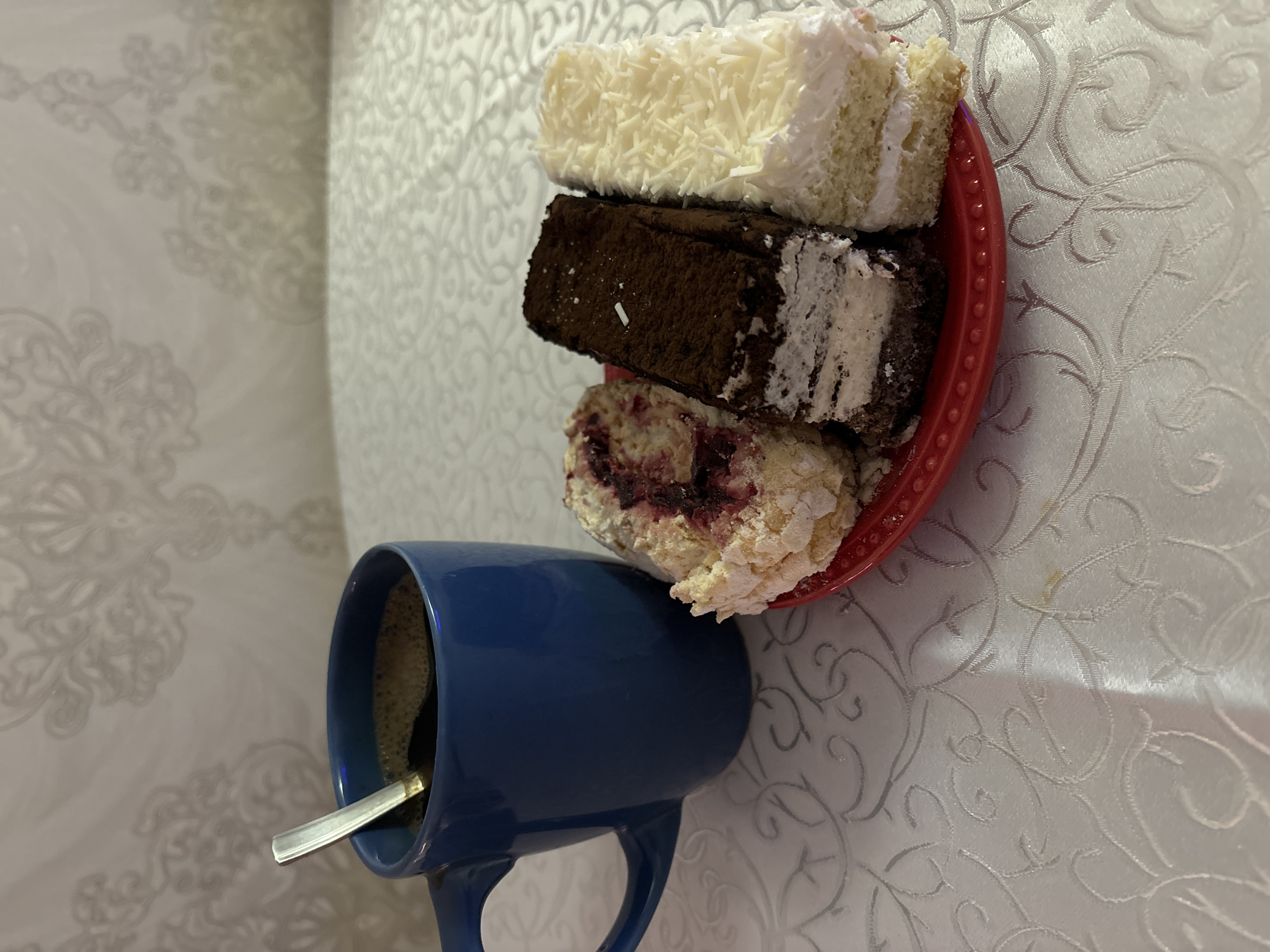 Here's a description of the image:
**Scene:** An indoor setting, likely a table surface covered with a textured, light-colored cloth.
**Objects:**
* A red plate stacked with three slices of cake. The top slice has white frosting and shredded white decoration. The middle slice is a chocolate-colored cake with some white frosting. The bottom slice has a light-colored frosting and some dark red or purple filling (possibly berries).
* A blue mug containing a dark liquid (likely coffee). A silver spoon is placed inside the mug.
**Location:** The city of Chaikovsky, Russia.