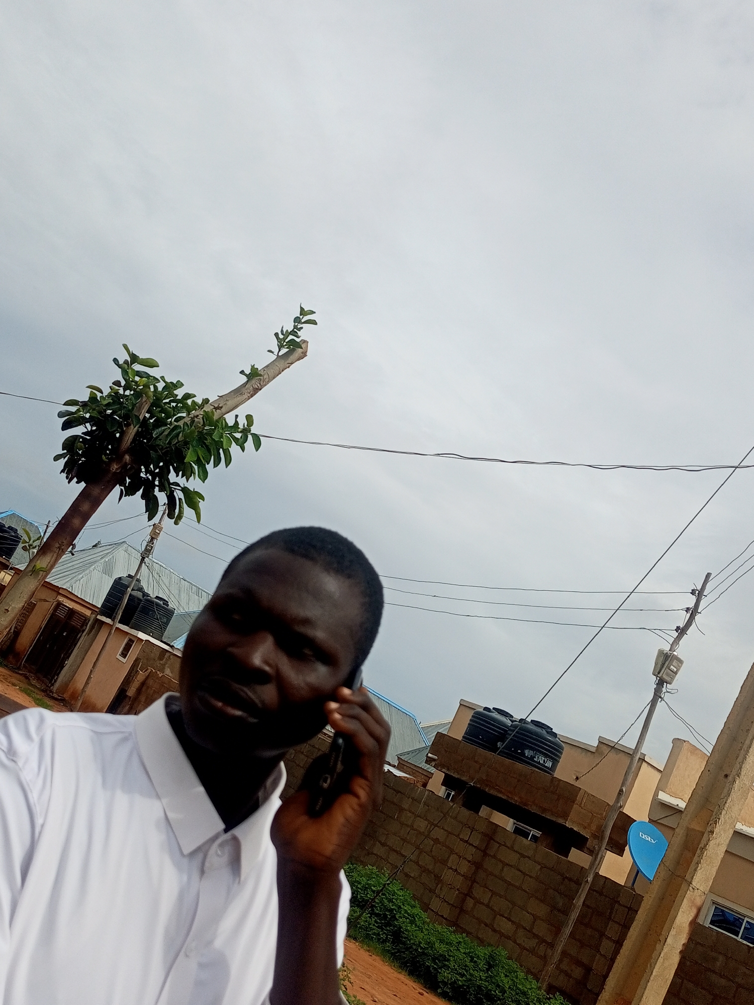 The image depicts a close-up of a young Black man wearing a white collared shirt and speaking on a mobile phone. He is positioned in the foreground, with his face partially obscured as he holds the phone to his ear. In the background, a tree with green leaves is visible, leaning at an angle. Behind the tree, several buildings with corrugated metal roofs and water tanks are present, along with a brick wall. Overhead, power lines crisscross the cloudy sky. A blue satellite dish with the "DSTV" logo is mounted on a pole to the right of the frame. The scene appears to be an outdoor urban or semi-urban environment.