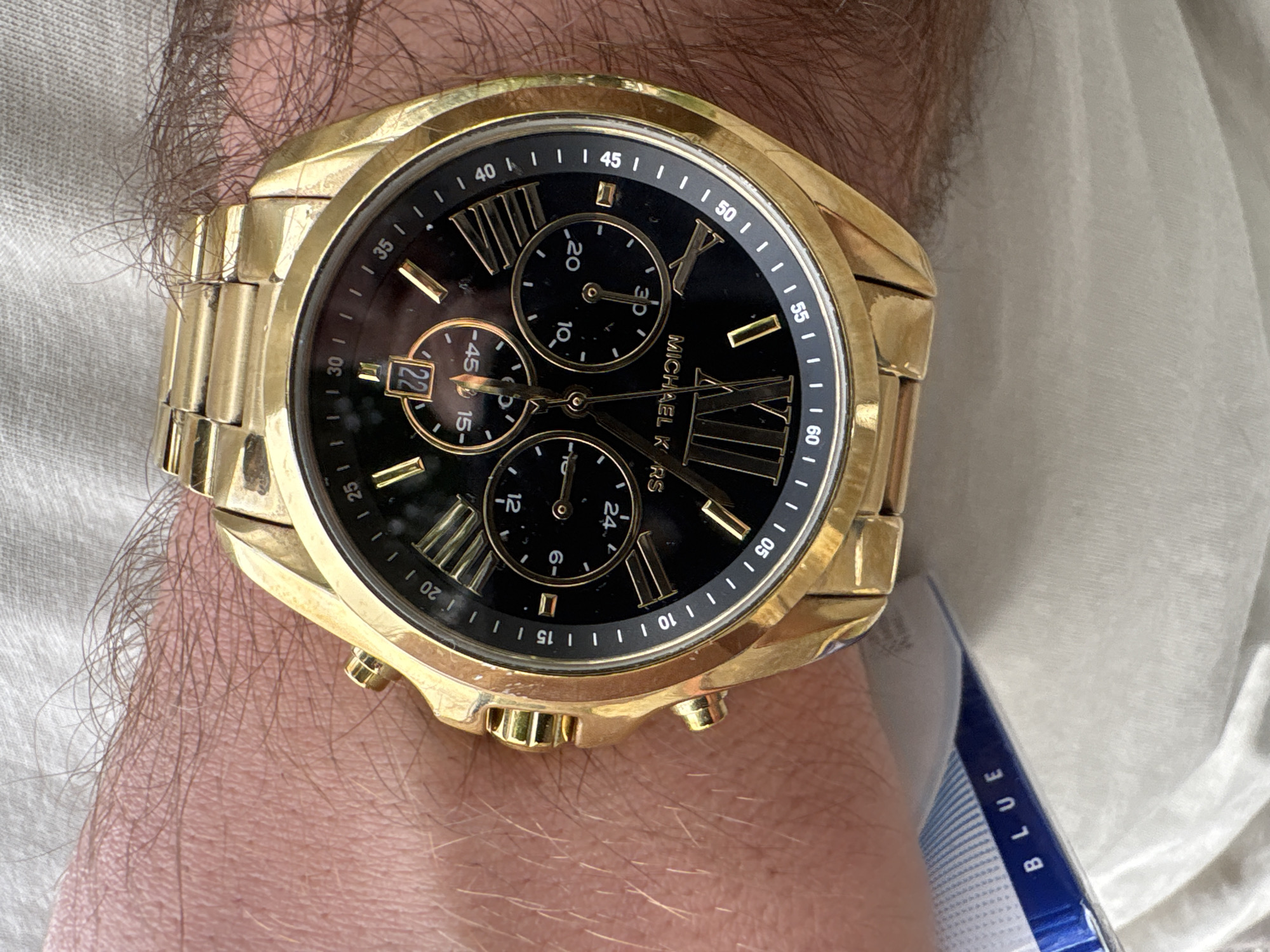 A close-up shot shows a Michael Kors gold wristwatch on a person's wrist. The watch has a black face with Roman numerals. The date is visible, displaying "22." The person has fair skin with arm hair. A white garment is in the background, along with a blue and white object with the word "BLUE" visible on it. The location is unknown and there is no indication that the photo was taken in Yerevan, Armenia.