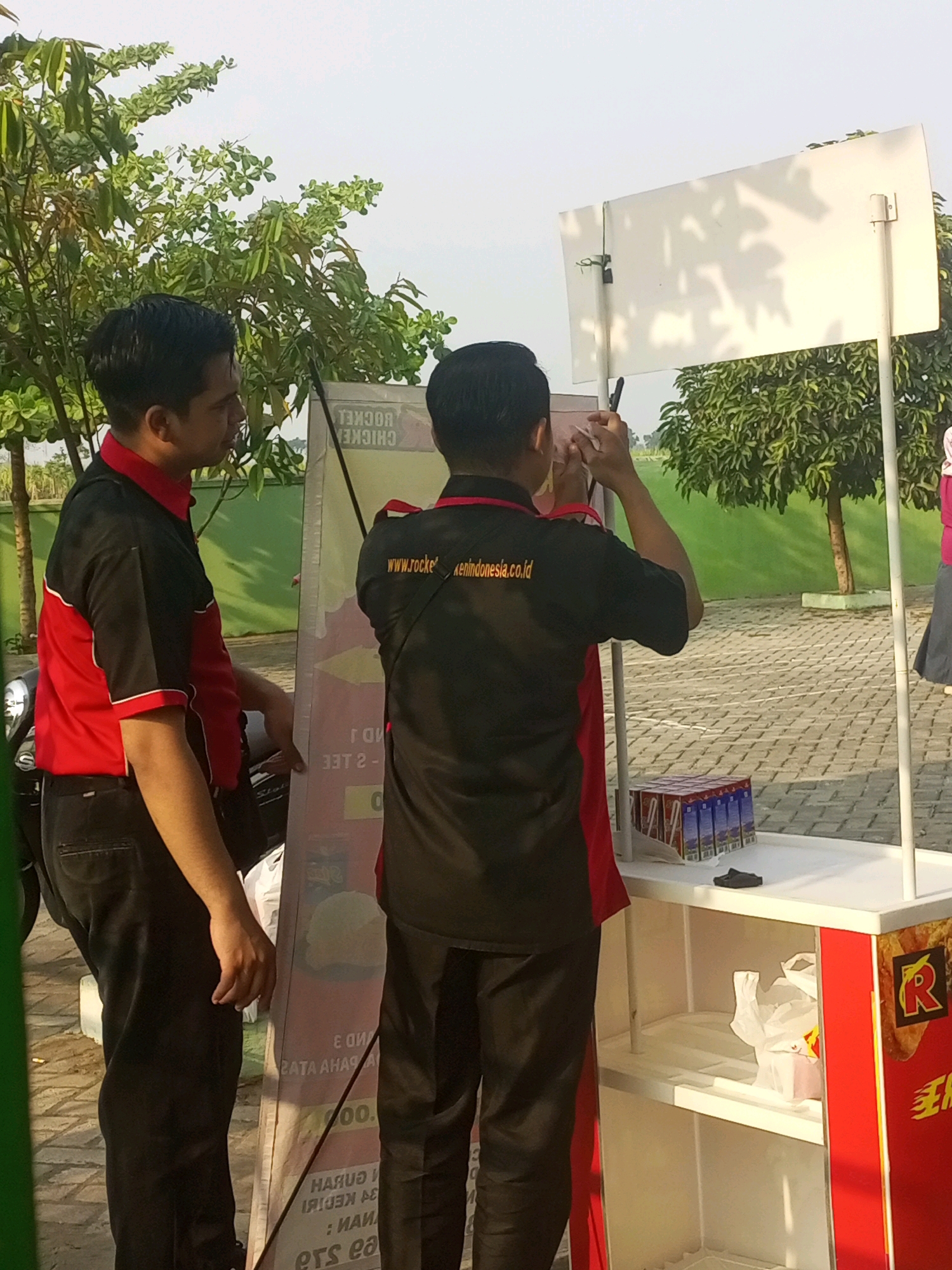 Two men, likely employees, stand near a food stall in Gurah, Indonesia. The man on the left wears a red and black uniform and is positioned facing the viewer, looking slightly to the right. He has dark hair and is wearing black pants. The man on the right, also in a black uniform with red accents, faces away from the viewer and is writing on a white board positioned on a pole. He also has dark hair.
The food stall itself is red and white, with a white countertop displaying several boxes of what appear to be drinks. The stall has open shelving underneath. A large, tall display board, partially visible, stands behind the man on the left. This board contains text and images, some of which are upside down. One visible text reads "www.rocketindonesia.co.id."
The environment is outdoors, with trees and foliage visible in the background. The ground appears to be paved with interlocking bricks. The lighting suggests it is daytime, possibly late afternoon, with shadows cast by the trees. The overall scene depicts a commercial setting, likely a promotional event or outdoor food vendor.