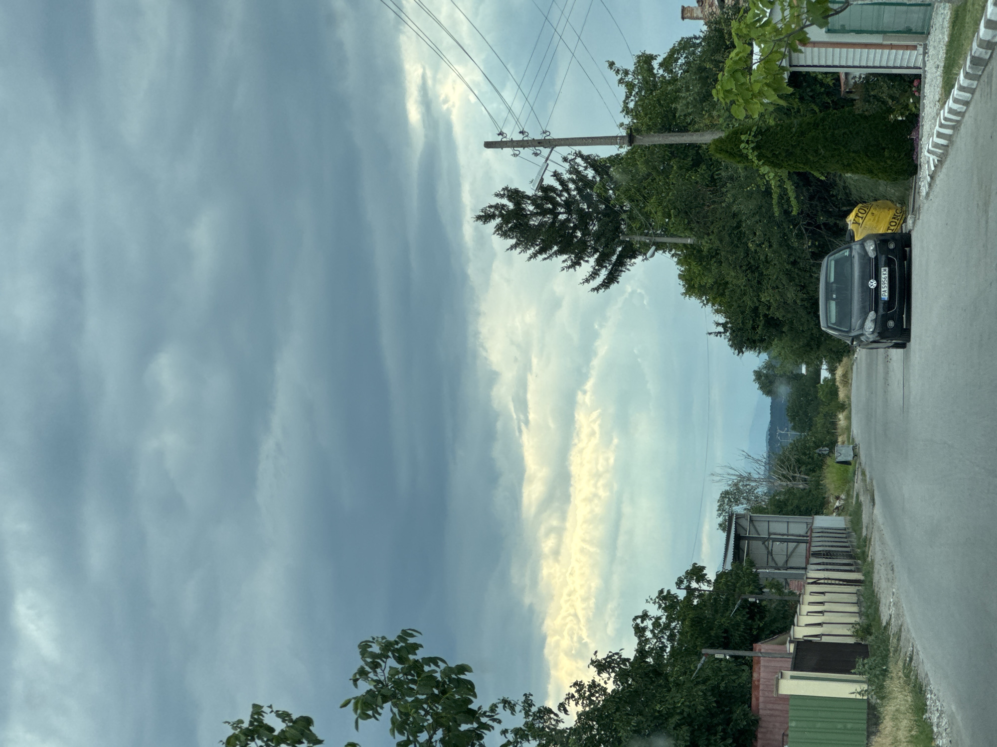 This is a vertical, outdoor photograph taken from a slightly elevated viewpoint, possibly from inside a vehicle. The scene depicts a rural road in Gelemenovo, Bulgaria, with buildings and lush greenery lining one side. The sky is a dominant feature, filled with dramatic, overcast clouds with streaks of bright sunlight breaking through in the distance, suggesting either the beginning or end of a storm, or perhaps a sunrise/sunset.
On the right side of the road, a dark-colored Volkswagen hatchback is parked. The car has a license plate visible, reading "PA 3956 KM". Next to the car, a bright yellow bag, possibly containing construction material, is visible with partial text that includes "YTONG" and "TONG". Further down the road, several simple, utilitarian structures with corrugated metal roofs are present. One structure appears to be an open-sided shed with metal shelving units inside. A power line pole with multiple wires stretches across the frame, adding to the rural atmosphere.
The vegetation is dense, with mature trees and bushes forming a green backdrop to the scene. In the far distance, hills or mountains are faintly visible under the cloudy sky. The overall mood is somewhat somber due to the overcast sky, but the patches of sunlight add a touch of hope or dramatic flair. The time of day is difficult to pinpoint precisely due to the cloud cover, but the quality of light suggests it might be late afternoon or early evening.