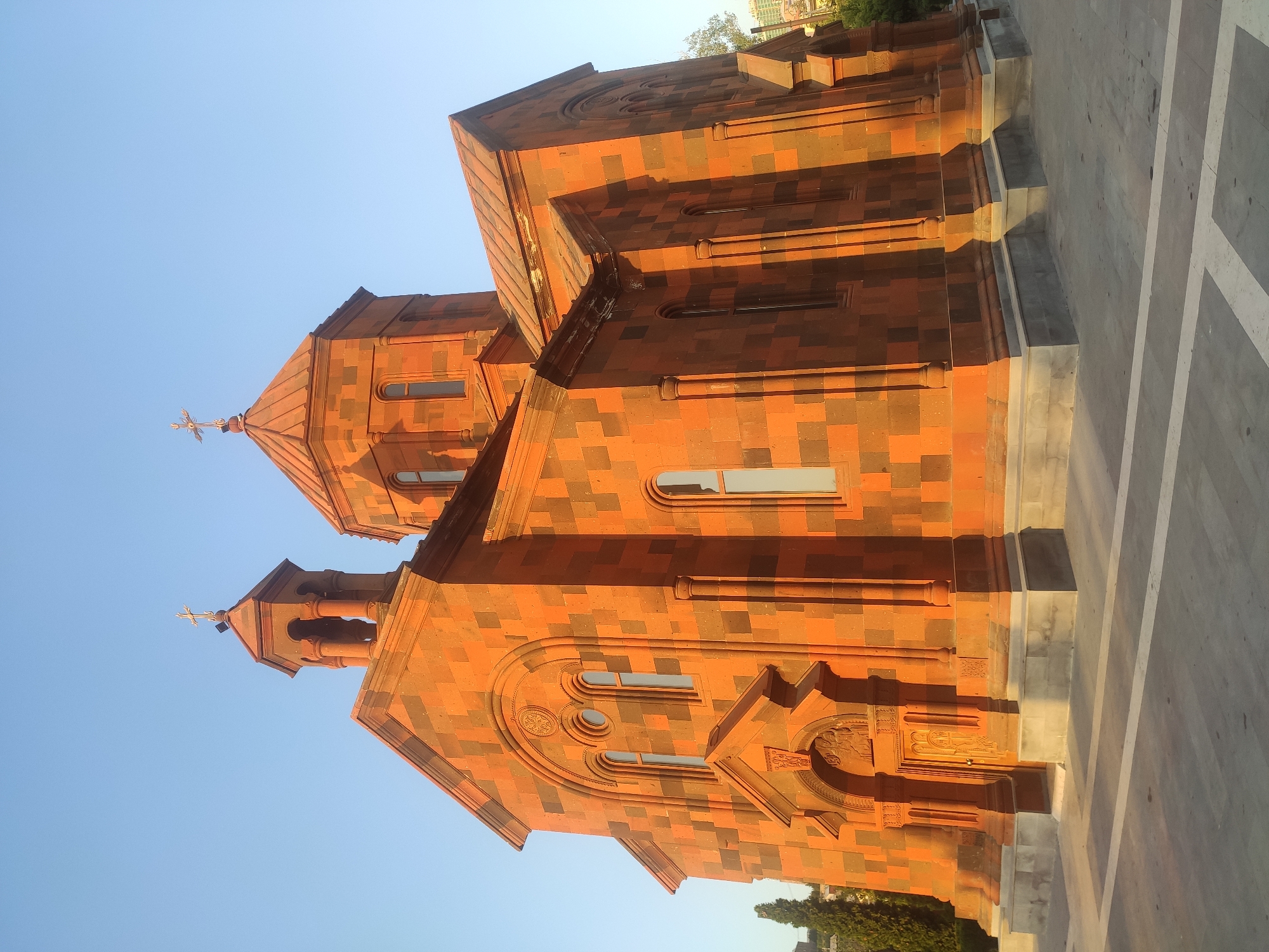 The image shows the Holy Trinity Church, a Catholic church located in the Kanaker-Zeytun administrative district of Yerevan, Armenia. The church is constructed from red and dark volcanic tuff, giving it a distinctive checkered appearance. It features a central dome topped with a cross, and a bell tower on the left side, also adorned with a cross. The church has arched windows and an ornate entrance. The building is illuminated by the warm light of the setting sun, casting long shadows on the paved plaza in front of it. A clear blue sky forms the backdrop. Green trees and a modern building are visible in the background on the right side of the image. There are no people present in the scene.