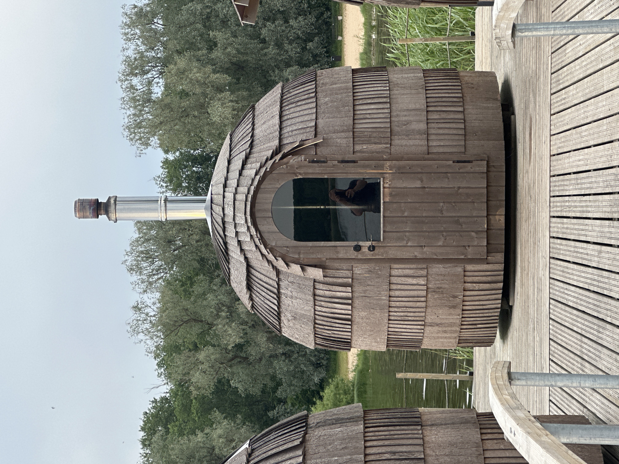 A cluster of three spherical wooden saunas with shingled roofs are situated on a wooden pier. The saunas are made of dark, weathered wood and have arched doorways with small windows. A metal chimney protrudes from the top of the central sauna. The pier overlooks a body of water, with lush green trees and a beach visible in the background. The sky is overcast. The scene is set in Tallinn, Estonia.