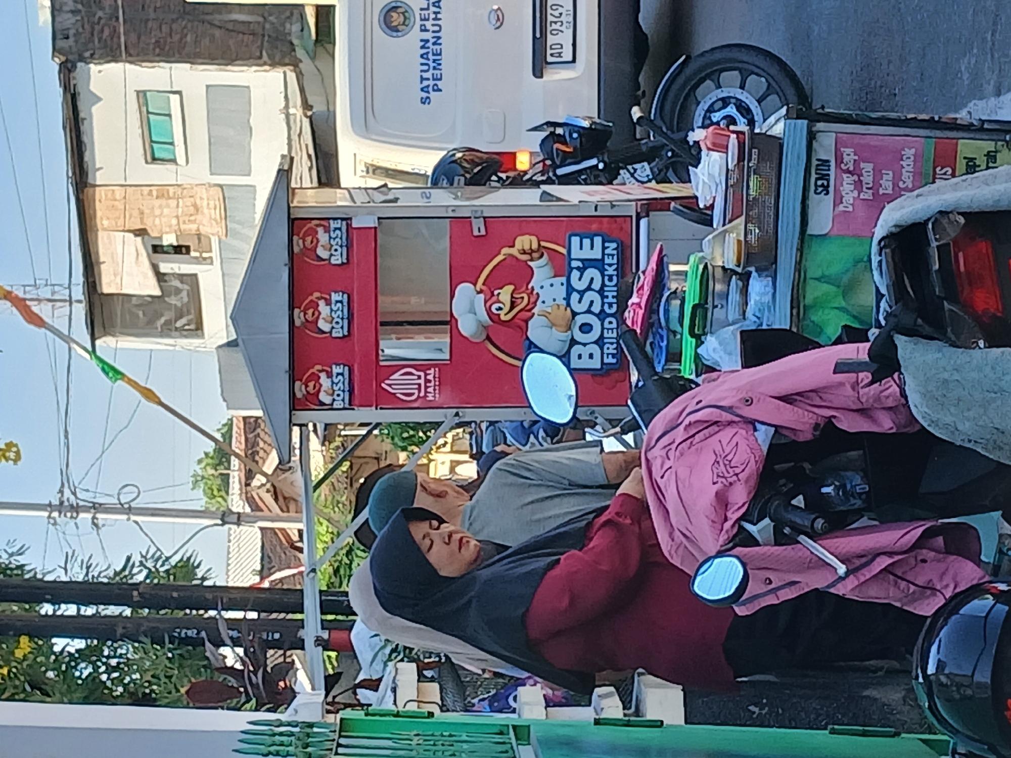 In Surakarta, Indonesia, on a sunny day, a woman wearing a black hijab and a maroon shirt stands in front of a food cart selling "Boss'e Fried Chicken." The cart features a cartoon chicken logo and the words "Halal" and "Boss'e Fried Chicken." Behind the cart, a white van with the text "Satuan Pelaksana Pemeliharaan" is parked. A motorcycle is parked next to the cart, with a pink jacket draped over its handlebars. In the background, there are buildings, trees, and power lines, suggesting an urban or suburban setting. Several other individuals are visible in the background, partially obscured by the foreground subjects. The scene appears to be a typical street-side food stall operation.
