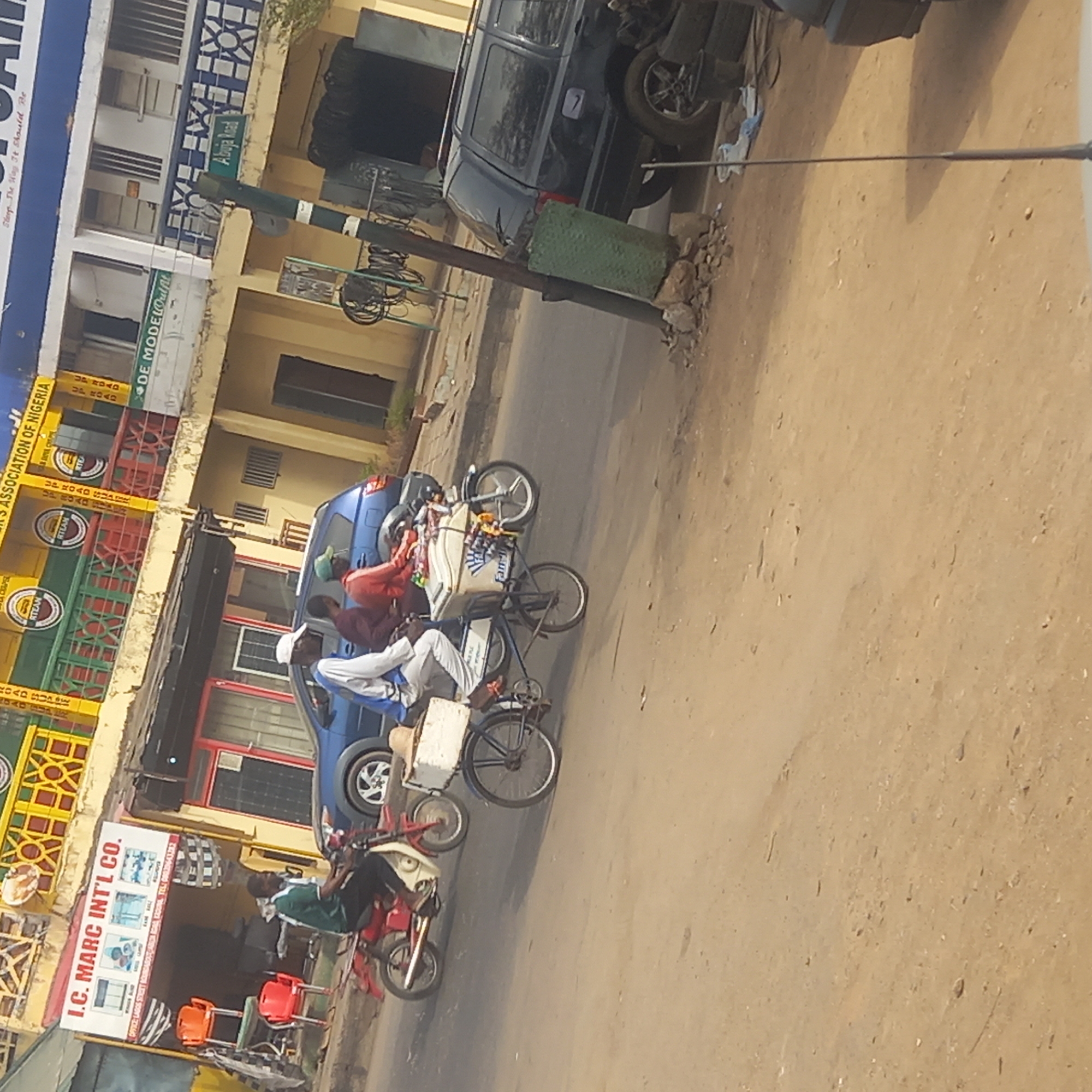This image captures a vibrant daytime street scene in what appears to be a commercial area, likely in Dadi Riba, Nigeria, given the provided context. The perspective is tilted, making the street run diagonally through the frame.
Several people are visible, engaged in various activities. In the mid-ground, a person rides a bicycle equipped with a large white cooler displaying the partial text "Fam," suggesting they might be a vendor selling beverages or snacks. Another individual sits on a red motorcycle nearby. Further up the street, a group of people are gathered around a blue car. A dark-colored SUV is parked further down the road, closer to the buildings.
The street itself appears to have both paved and unpaved sections, with patches of dirt or sand visible. Buildings line one side of the street, featuring different architectural styles and vibrant colors.
Notable details include several business signs. A prominent building is decorated in yellow and green with the repeated acronym "RTEAN," identifying it as associated with the "ROAD TRANSPORT EMPLOYERS' ASSOCIATION OF NIGERIA." Another sign reads "I.C. MARC INT'L CO." with an address including "LAGOS STREET BOUNDARY AREA KADUNA, KADUNA," and a phone number. A street sign clearly indicates "Abuja Road." Other partial texts like "DE MODELOulfit" and "Sleep The Way It Should Be" (possibly a furniture or bedding store) are also visible. The scene depicts typical daily street life in a bustling urban or semi-urban environment.