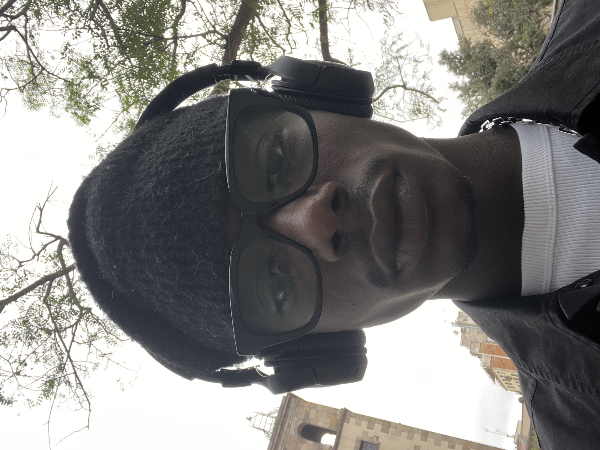 A low-angle, close-up photograph depicts a person wearing a black beanie, large dark-rimmed glasses, and black over-ear headphones. The individual is also wearing a white ribbed undershirt and a dark jacket with a silver chain visible at the neckline. The person's face is in sharp focus, revealing their skin tone, facial features, and reflections of the surroundings in their glasses. Behind the subject, tree branches with sparse foliage are visible against a bright sky. A portion of a beige stone building with architectural details is visible in the lower part of the background. The building's style, the foliage, and overall aesthetic suggest the location is likely Barcelona, Spain.