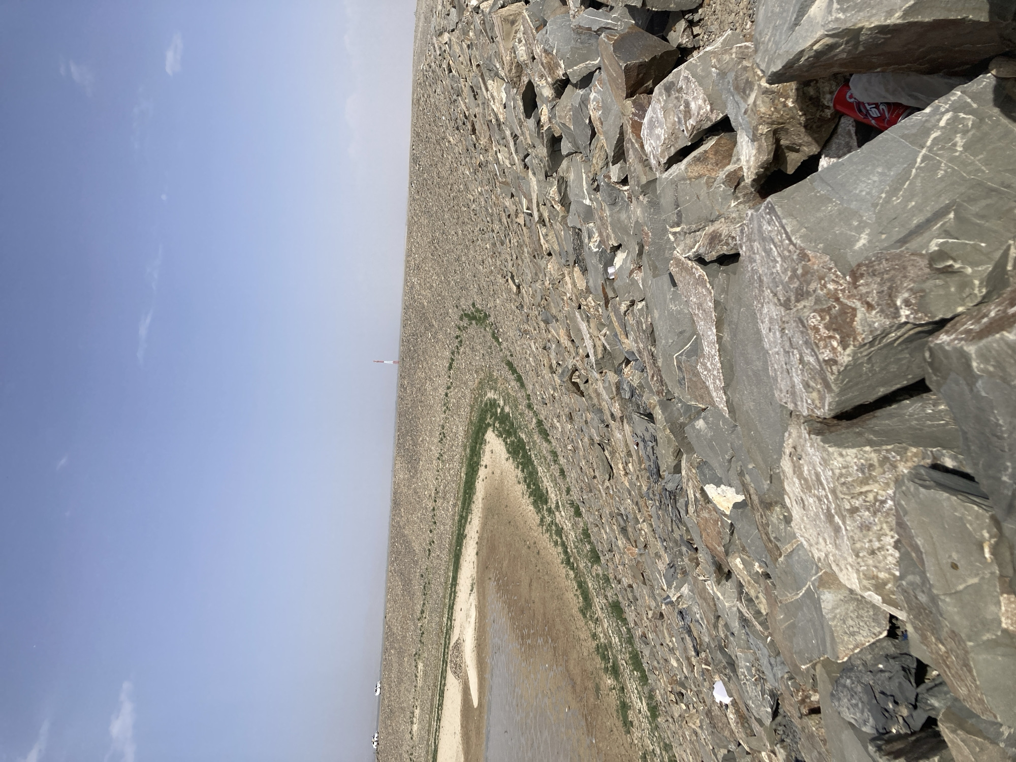 The image shows a wide, man-made embankment or dam composed of large, angular rocks, with a body of water (possibly a reservoir or river) in the distance, located in Gulistan, Pakistan. The sky is a pale blue with some haze. A possible radio tower is faintly visible in the distance. A discarded soda can is wedged between rocks. Small white spots near the body of water. No people are visible. The embankment is densely packed with rocks of varying sizes and shades of gray and brown. A line of greenery traces the edge of the water body. The orientation of the image is on its side, making the embankment appear to rise vertically.