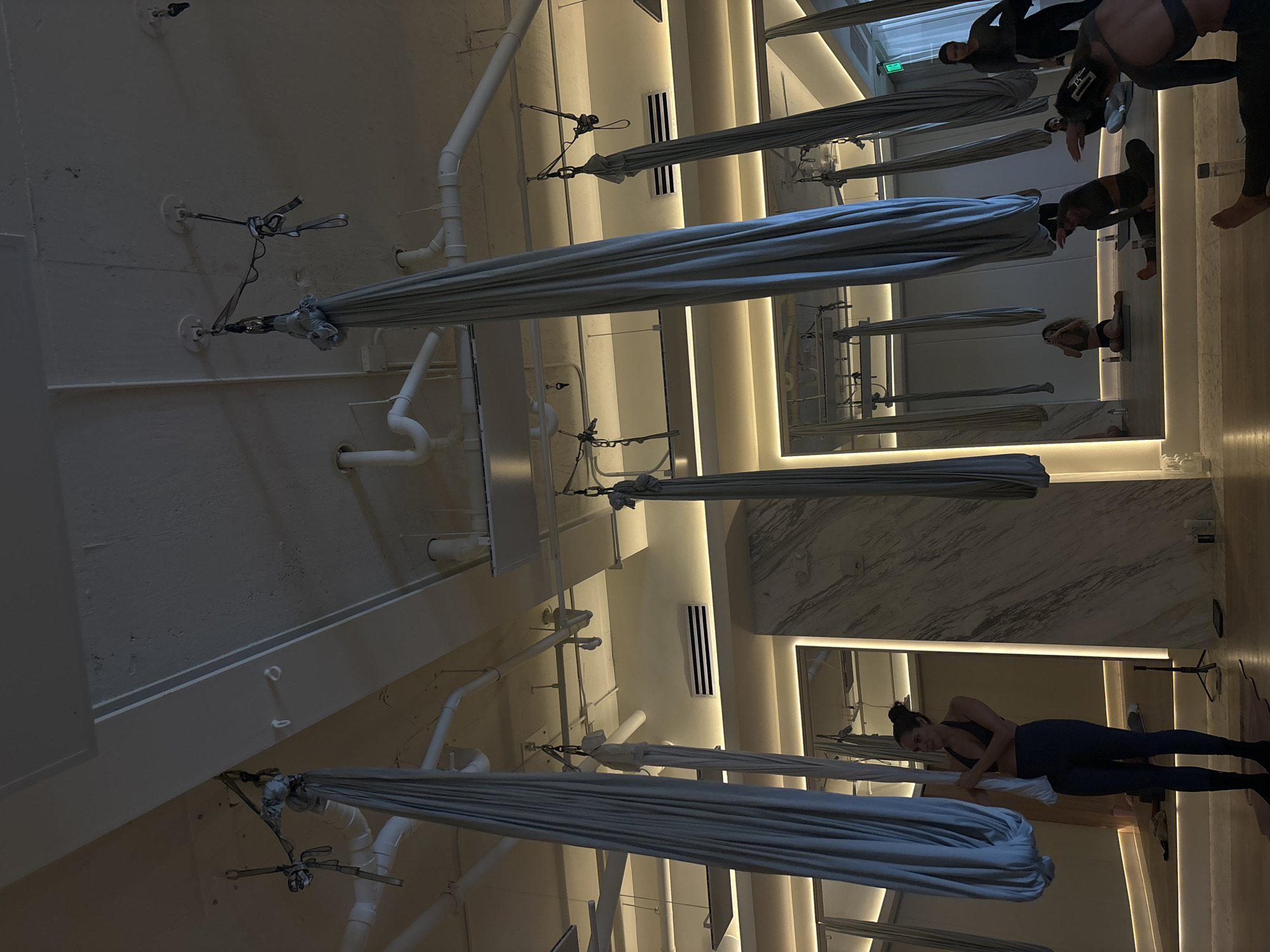 The image captures the interior of an aerial yoga studio, likely located in Miami, United States of America. The studio features several aerial silks suspended from the ceiling. The ceiling itself is white with exposed pipes and support structures. There are also ventilation grilles and recessed lighting fixtures. The walls appear to be white or light-colored.
Several individuals are present in the studio, some of whom are engaged in aerial yoga poses or activities. There are mirrors on the walls.