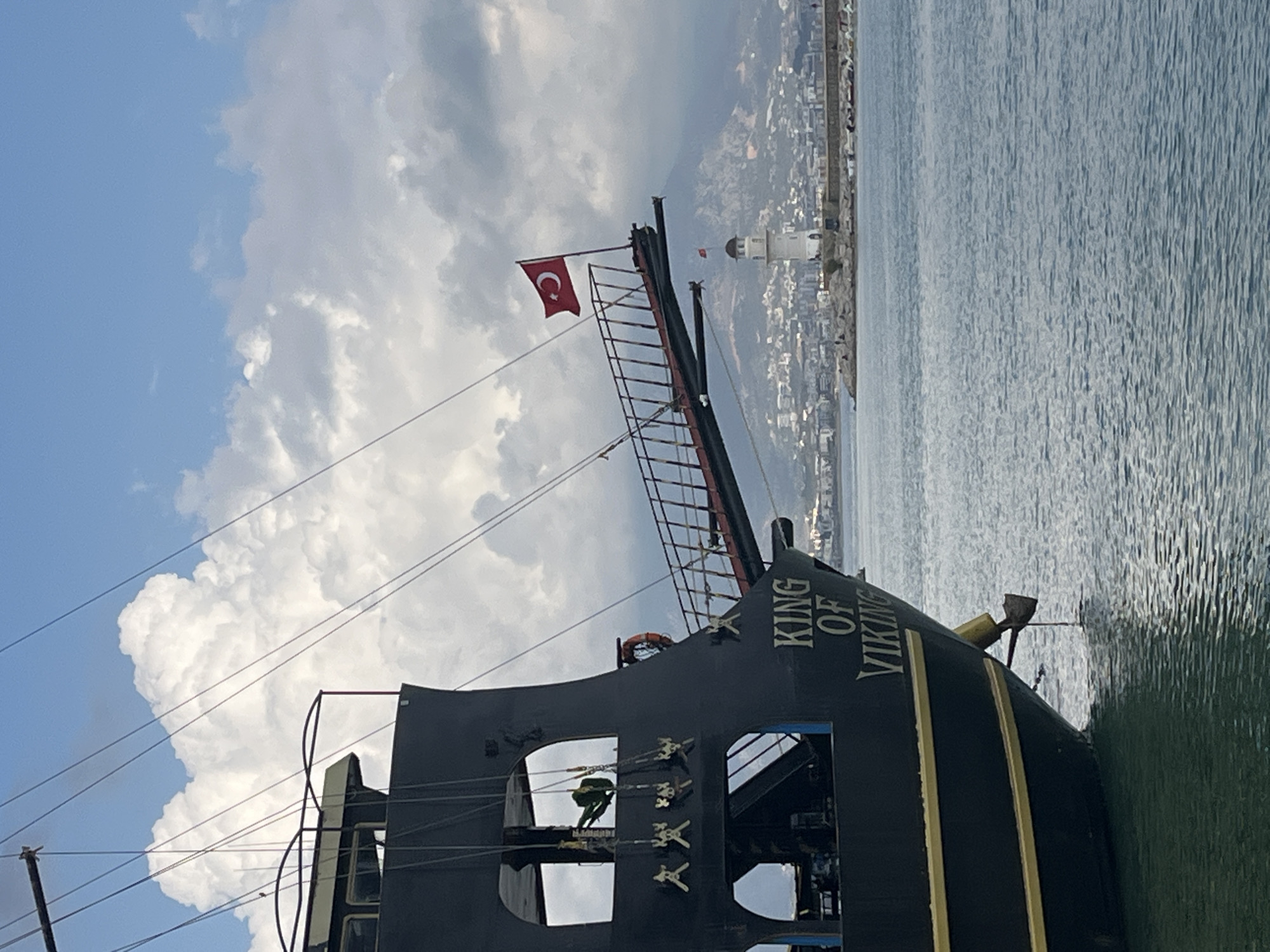 A daytime, medium shot depicts a docked pirate ship named "King of Viking" in Alanya, Turkey, against a backdrop of cloudy skies and calm water. The Turkish flag flies atop the ship's mast. In the distance, a white lighthouse stands among the buildings of Alanya. The ship, predominantly black with gold accents, features a visible ladder leading up the mast. Skulls decorate the ship's exterior. No people are visible on deck.