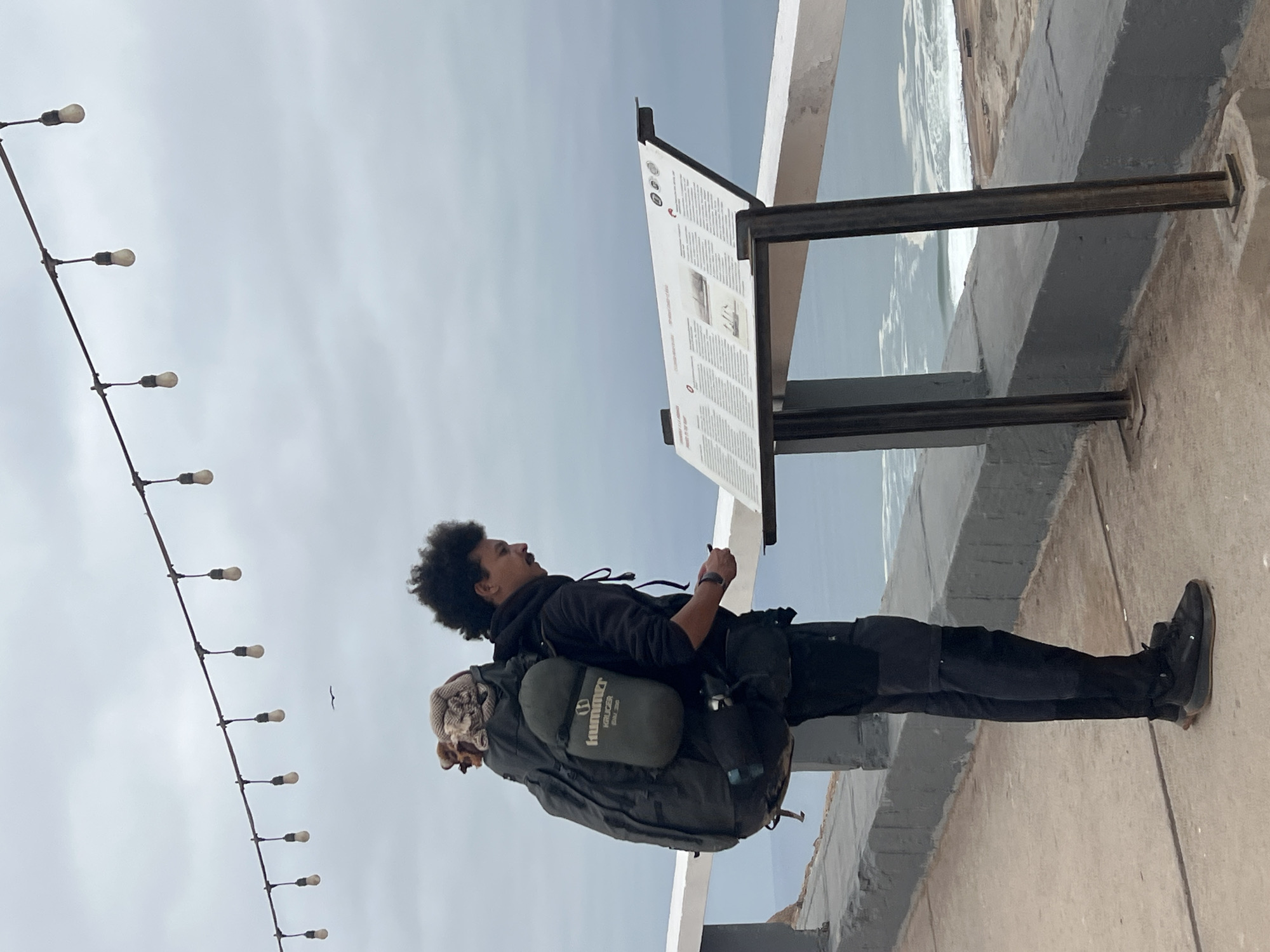 An outdoor scene depicts a male individual standing on a concrete walkway beside a low gray wall topped with a white railing, overlooking the Pacific Ocean under an overcast sky. The person, positioned in profile facing right, is a young adult male with dark, curly hair and a mustache. He wears a dark hooded sweatshirt, dark cargo-style pants, and dark athletic shoes with brown soles. He carries a large, dark-colored hiking backpack with a light-colored, patterned item strapped to the top and a green sleeping bag, labeled "Hummer KRUGER BMV 200," attached to its side. His left hand holds a small dark object at waist height, and his right hand is slightly raised. He is engaged in viewing a white information sign, angled towards him, which is mounted on two dark metal posts. The sign displays multiple columns of text, likely in Spanish, and features small circular icons at the top right, with a small image in the center. In the background, white waves break in the grayish-blue ocean, with what appears to be a sandy or rocky shoreline visible further out. Overhead, a string of nine visible incandescent light bulbs hangs from a wire stretched across the upper frame. A small dark bird is in flight against the cloudy sky, below the string lights. The scene is consistent with a coastal viewpoint in Arica, Chile.