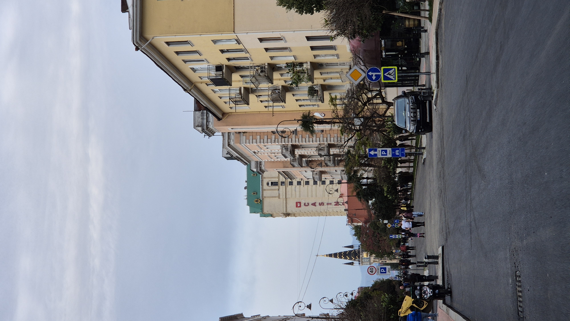 This is an eye-level outdoor shot of a street in Batumi, Georgia. The camera is oriented parallel to the street, with the view looking down its length. The sky above is overcast with thin white clouds.
On the right side of the frame is the asphalt street. A dark-colored SUV is parked near the sidewalk. Several street signs, including parking signs and a pedestrian crossing sign, are visible.
On the left side of the frame are buildings. The buildings closest to the camera are multi-storied and yellow with ornate balconies. Further down the street, a building with a green roof and a sign that reads "CASINO" can be seen. A distinctive architectural tower with dark spires is visible further down the street.
Several people can be seen walking on the sidewalk. Some are individuals, while others are in small groups. Near the bottom of the frame are parked motorcycles and bicycles. Trees with bare branches line the street, interspersed with ornate street lamps.