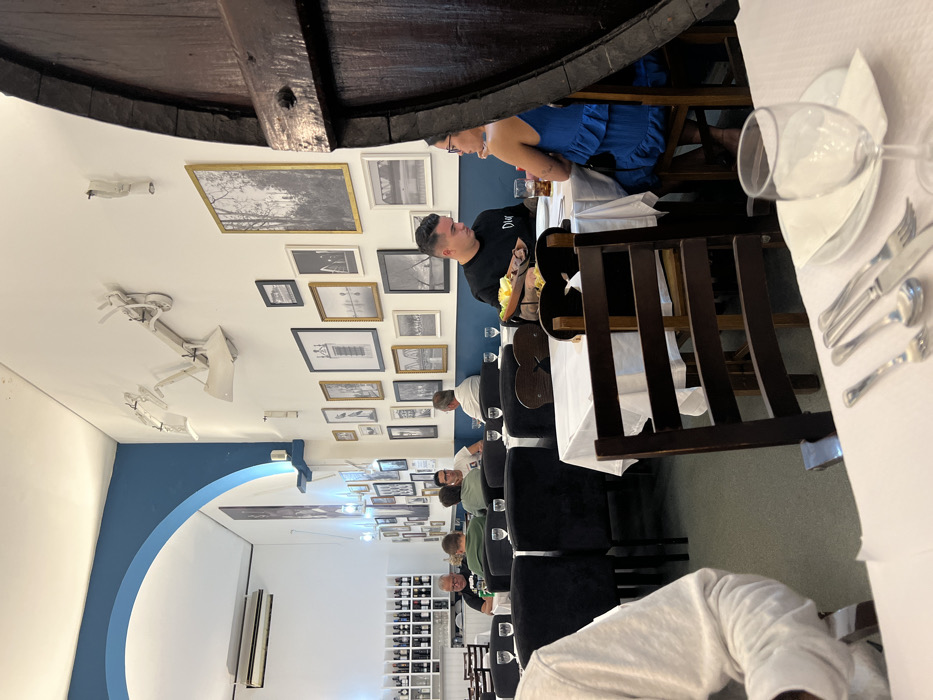 The image depicts the interior of a restaurant in Coruche, Portugal. The scene is a well-lit dining area with white walls and a distinct blue trim outlining an archway and a lower wall section. A collection of framed monochrome photographs of various sizes adorns the main wall, complemented by abstract white sculptural elements mounted higher up.
Multiple dining tables are present, each covered with a white tablecloth. Chairs are a mix of dark upholstered models and dark wooden styles. In the mid-ground, a man with short dark hair, wearing a black t-shirt with a white logo, is seated at a table opposite a woman with dark hair, wearing a blue tiered dress. A basket containing food, along with drinks in glasses, rests on their table. They appear to be engaged in conversation. Partially obscuring the right side of the frame is a large, dark-stained wooden barrel.
Further into the background, several other patrons, predominantly men, are seated at tables, some appearing to be conversing or dining. One man wears a green shirt, another a white shirt. On the far left in the background, a white shelving unit is stocked with numerous wine bottles. An air conditioning unit is mounted high on the wall near the archway. In the immediate foreground on the left, a portion of a person wearing a white long-sleeved shirt is visible. A table setting in the foreground displays silverware (fork, spoon, knife), a white plate, a white napkin, and an empty stemmed glass.