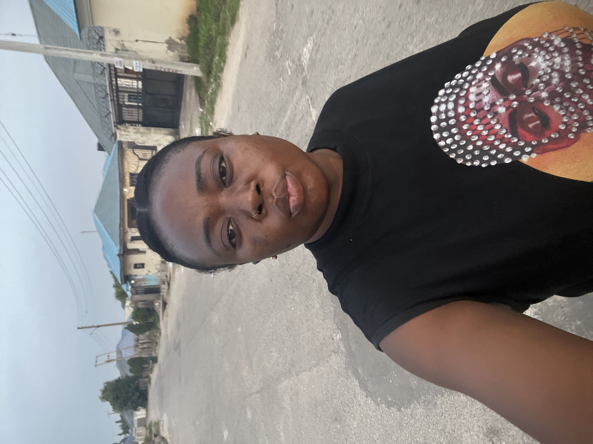 A medium close-up captures a person wearing a black T-shirt with a prominent image adorned with white gemstones. The person has a nose ring and is positioned at an angle, with a view down a street visible behind them. The street seems to be located in a residential area of Abuja, Nigeria, based on the architectural style of the buildings and the environment. There is pavement, and a few buildings with blue and gray roofs can be seen. The sky appears overcast or hazy.