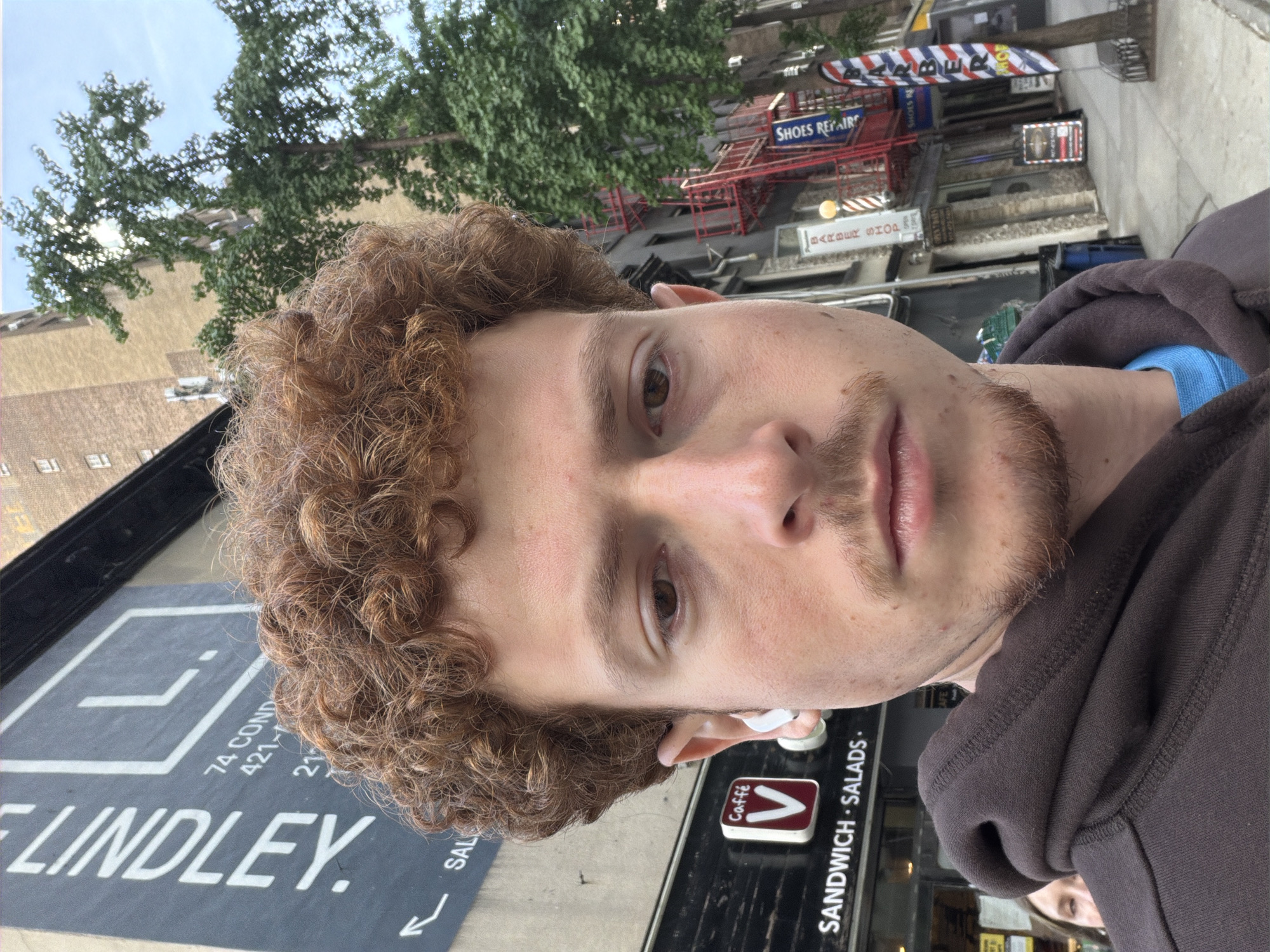 The image is a close-up shot of a man with curly hair, wearing a brown hoodie with a blue undershirt, and earbuds. He is standing in front of "Caffé V," a cafe advertising "Sandwich Salads." Adjacent is signage for "74 Cond" and "Lindley." A "Shoes Repair" shop is nearby, along with a "Barber Shop" with a barber pole. The image was taken in New York City, United States of America.