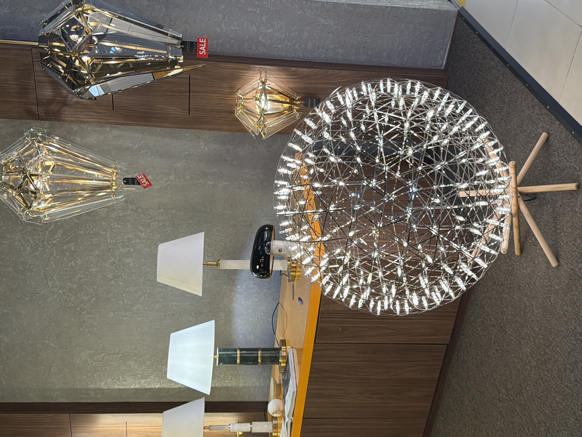 The image shows a well-lit interior space, likely a retail store, displaying a collection of modern lighting fixtures. In the foreground, a large spherical floor lamp with numerous small lights is prominently featured, supported by a wooden tripod base. Behind this, on a reflective orange surface, are three table lamps with white shades, one of which has a dark, curved base. Further back, two decorative pendant lamps, shaped like faceted diamonds, hang from the ceiling. Red "SALE" tags are visible on the pendant lamps, indicating a promotional event. The background features a textured gray wall and wooden paneling, suggesting a sophisticated and contemporary design. The overall impression is that of a showroom designed to showcase high-end lighting products. No people are visible in the image. The location is identified as Moscow, Russia.