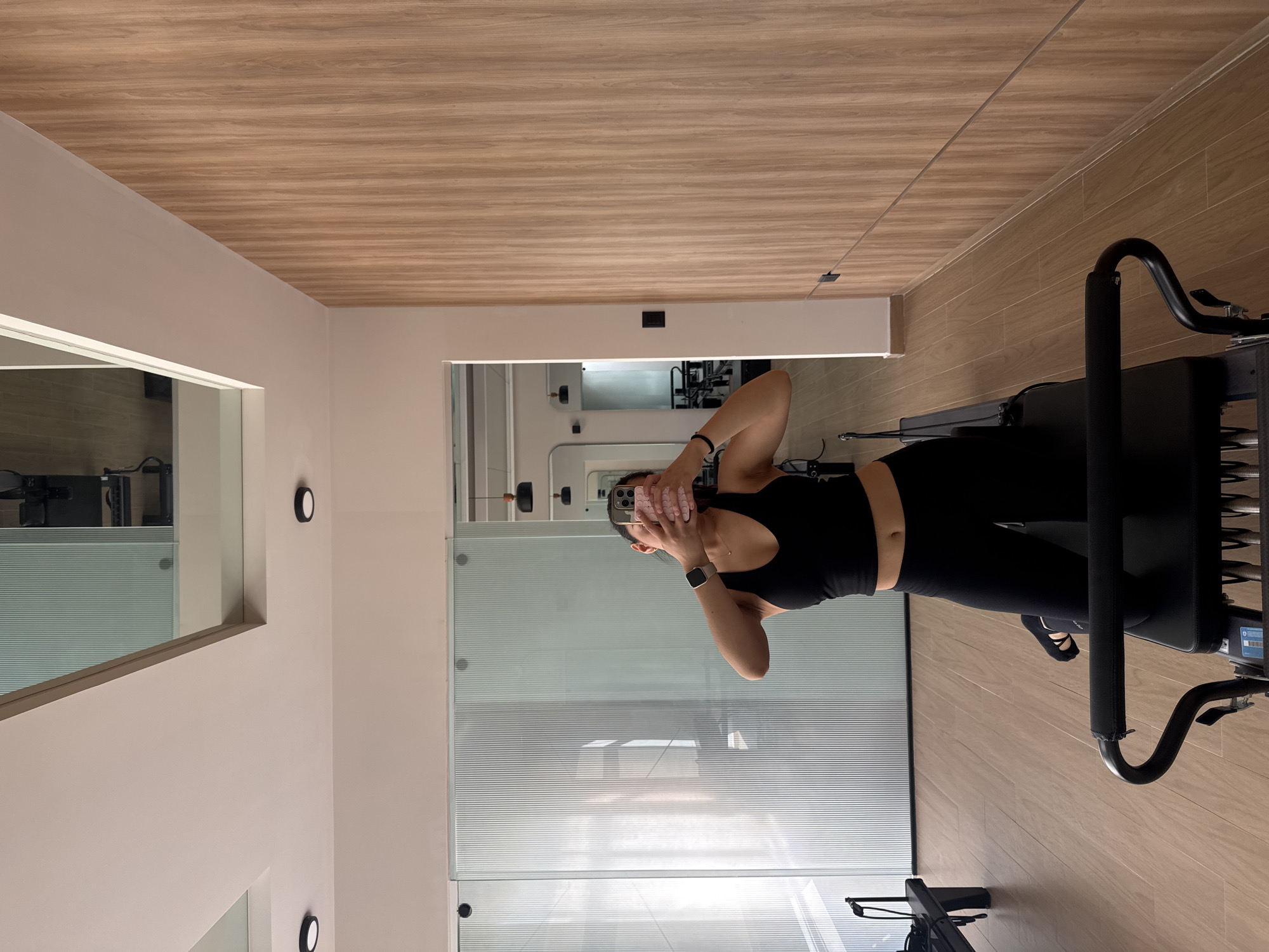 An indoor scene depicts a person in a modern fitness studio or gym, located in San Bernardino Tlaxcalancingo, Mexico. The image is oriented sideways, but viewed correctly, a person, likely female, stands upright towards the right side of the frame. She is wearing black activewear, including a sports bra that reveals her midriff and full-length leggings. She holds a light-colored smartphone with a textured case in front of her face, suggesting she is taking a mirror selfie or recording. A smartwatch is visible on her left wrist, and a dark band on her right.
To her right, a large piece of black exercise equipment, possibly a Pilates reformer, is visible, extending out of the frame. The studio features light beige walls and a distinctive aesthetic with light brown wooden plank flooring that extends up to cover the ceiling. A ceiling bulkhead with a light-colored soffit runs across the top part of the image.
Behind the person, there's a wall or partition made of frosted or textured glass with horizontal lines, reflecting parts of the room. On a cream-colored wall to the left, two circular wall-mounted lights with dark rims and white centers are visible. The overall setting appears clean and well-lit, characteristic of a contemporary fitness environment. No discernible text is clearly legible on any objects or surfaces. The activity is centered around a personal fitness session or a photographic moment within the studio.