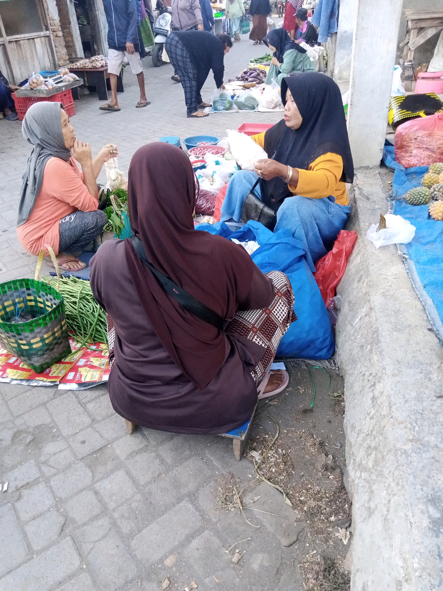 A street market scene is depicted in Sono Ageng, Indonesia, featuring several individuals engaged in commerce. Two women, both wearing hijabs, are the central figures. One woman, wearing an orange shirt and gray hijab, is seated and looking towards the left, her right hand touching her chin. She appears to be conversing with the other woman. The second woman, dressed in a yellow long-sleeved shirt and black hijab, is seated and holding a white plastic bag, seemingly engaged in a transaction or preparation of goods. A third person, also in a hijab and a brown robe, is seated in the foreground, facing away from the camera.
The ground is paved with interlocking gray bricks, with patches of dirt and debris. Various goods are displayed on the ground, including long green beans in a woven green and yellow basket, and several blue plastic bags and a red plastic bag. In the background, other market-goers and stalls are visible, including a stall with produce and a white scooter. The overall impression is of a busy, local market environment.