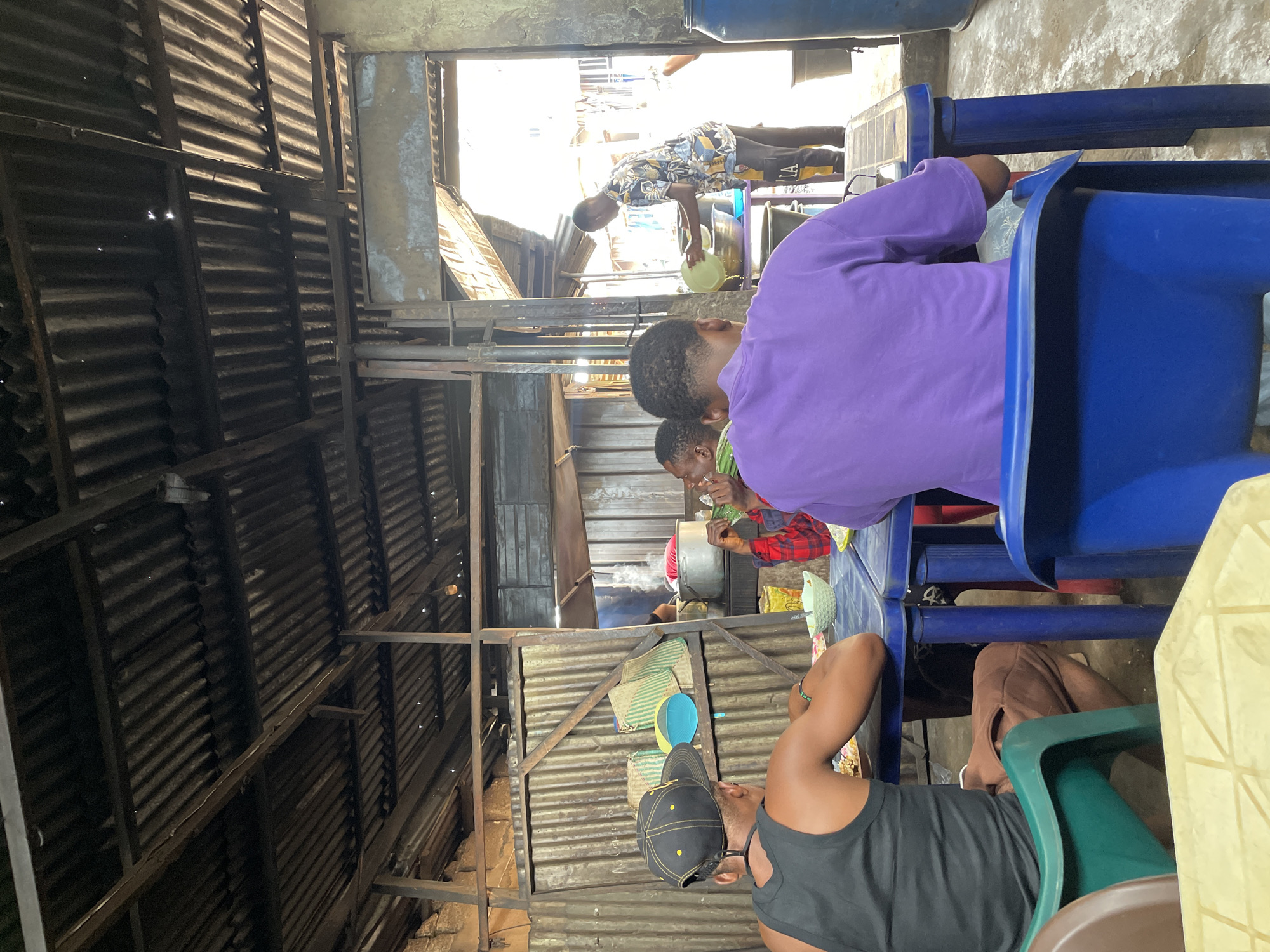 The photo shows an interior scene in Onitsha, Nigeria. Four people are visible, seated at blue and green plastic tables. The building appears to be constructed from corrugated metal sheets. A young boy wearing a patterned shirt is visible through a doorway, holding a green bowl. People in the foreground are wearing a black shirt, and purple long sleeve shirt. The environment seems to be in a market or informal setting.