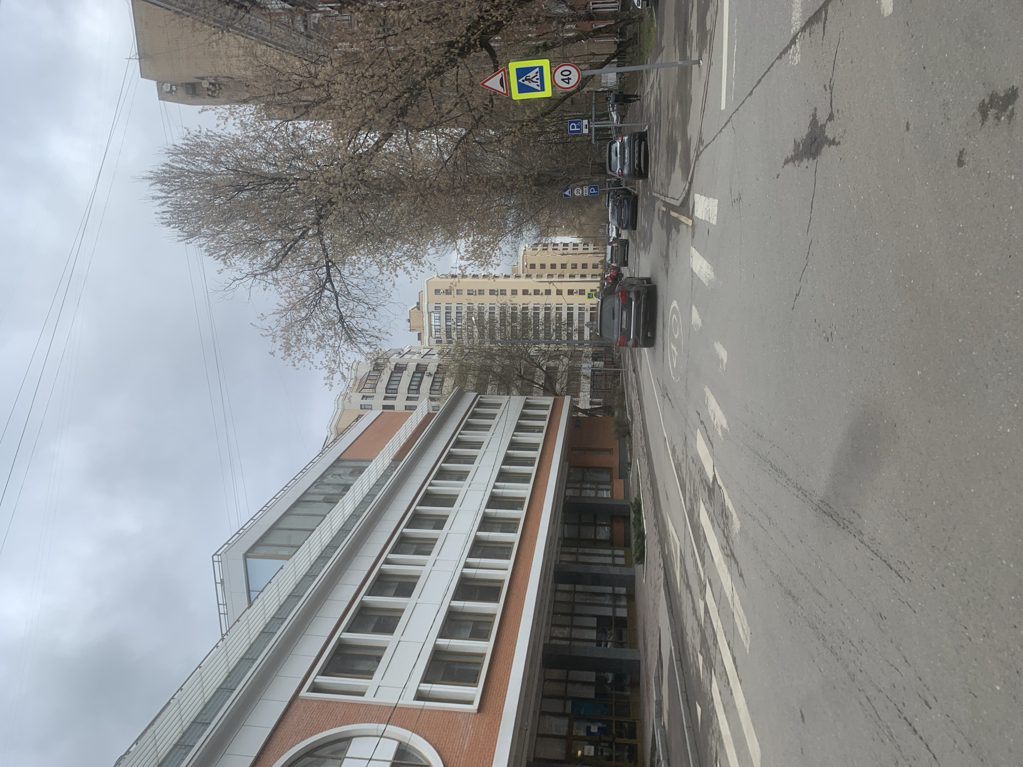The image shows a street scene in Moscow, Russia, on an overcast day. A modern, multi-story building with a distinctive facade of brick and white paneling dominates the left side of the frame. The street is wet, suggesting recent rainfall. Several cars are parked along the right side of the road, with others driving in the opposite direction. A pedestrian crossing is visible. Street signs indicating a pedestrian zone and a speed limit of 40 km/h are present. Bare trees line the street. In the background, taller apartment buildings are visible. No people are clearly identifiable in the image. The scene is urban and appears to be residential or commercial.